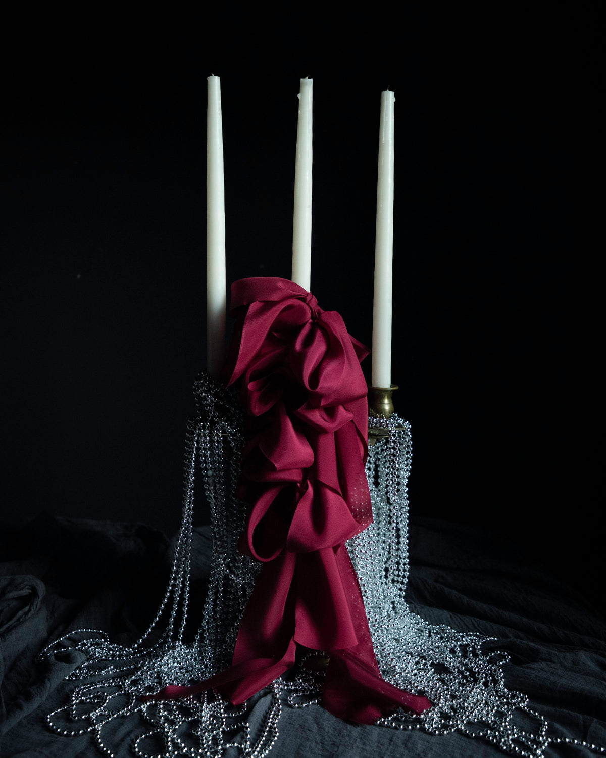 A gothic-style three taper candle holder with white candles, a large dark burgundy satin bow, and cascading silver bead chains on a black fabric background.