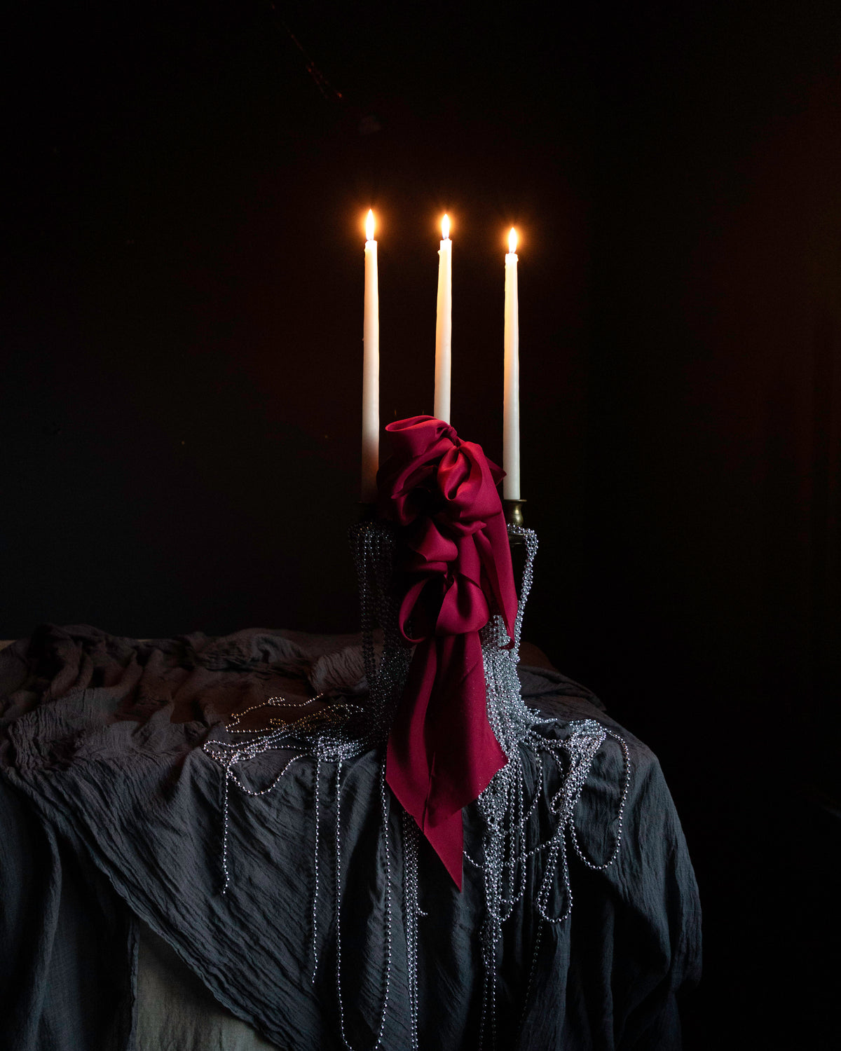 candles draped with metal beads and red ribbon bows on layered gray table textiles against a dark background