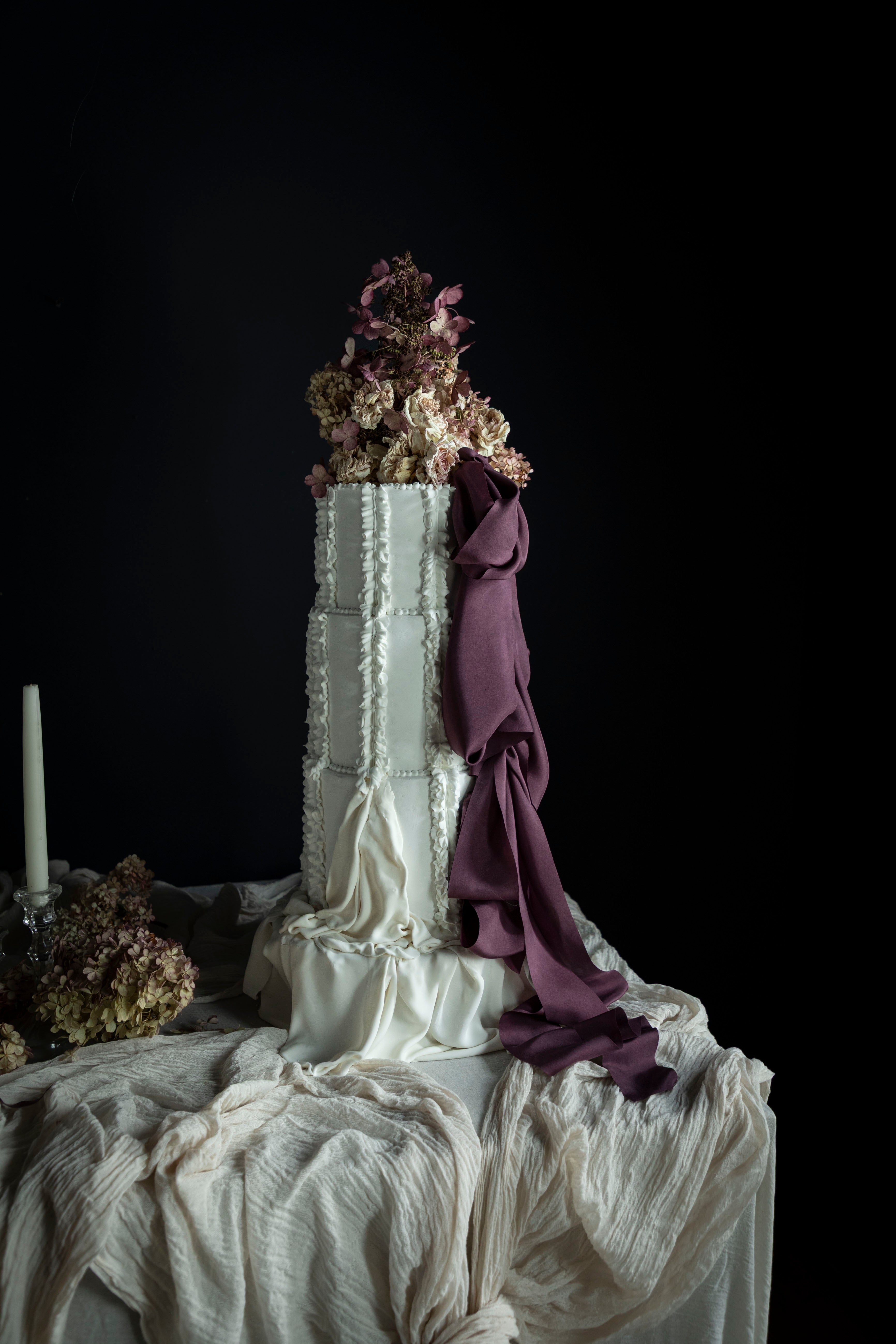 Decorative cake with white frosting, purple silk ribbon, and floral elements on a dark background