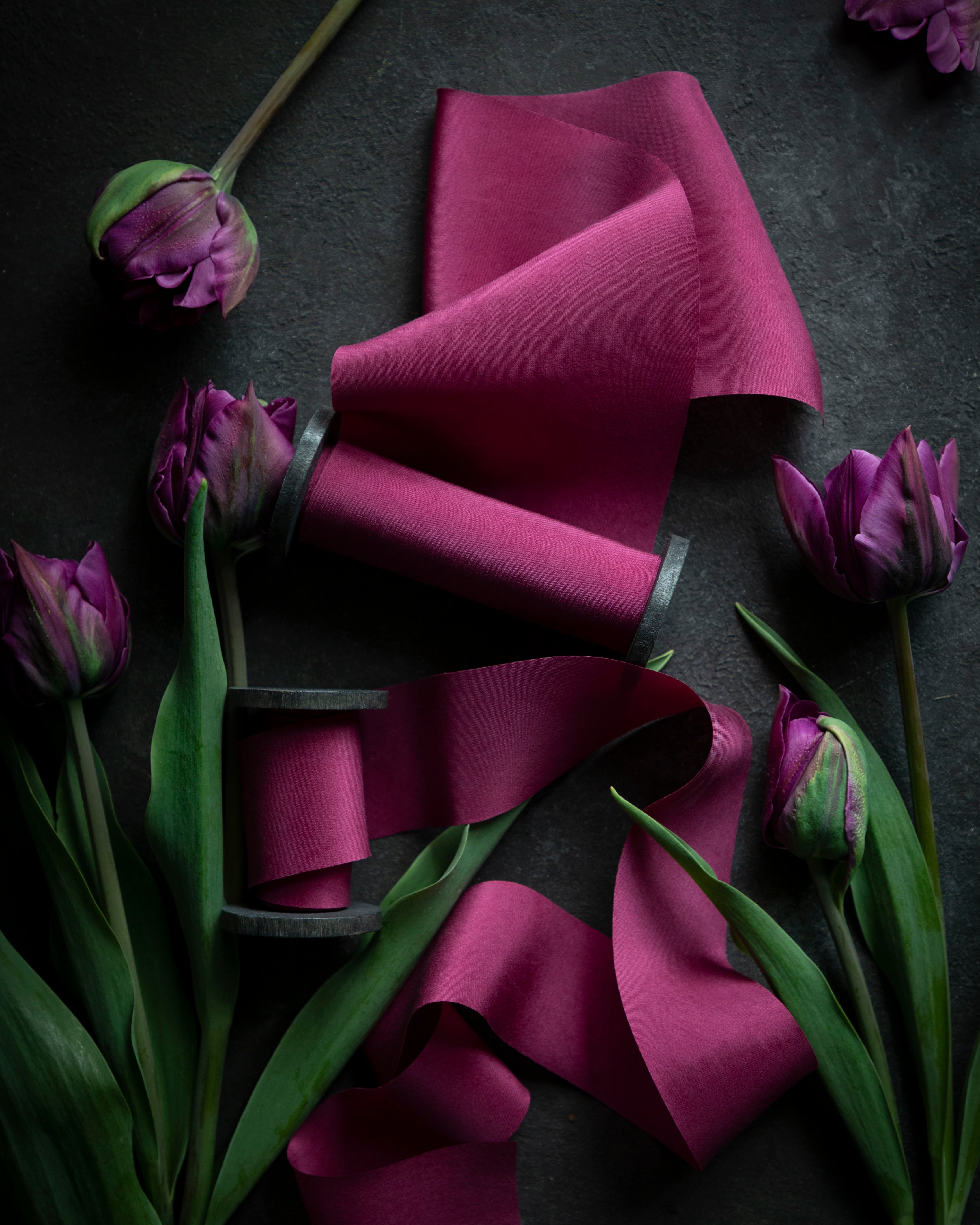 A series of vibrant pink silk ribbons arranged on a dark surface, surrounded by purple tulips, emphasizing the color and texture of the ribbons.
