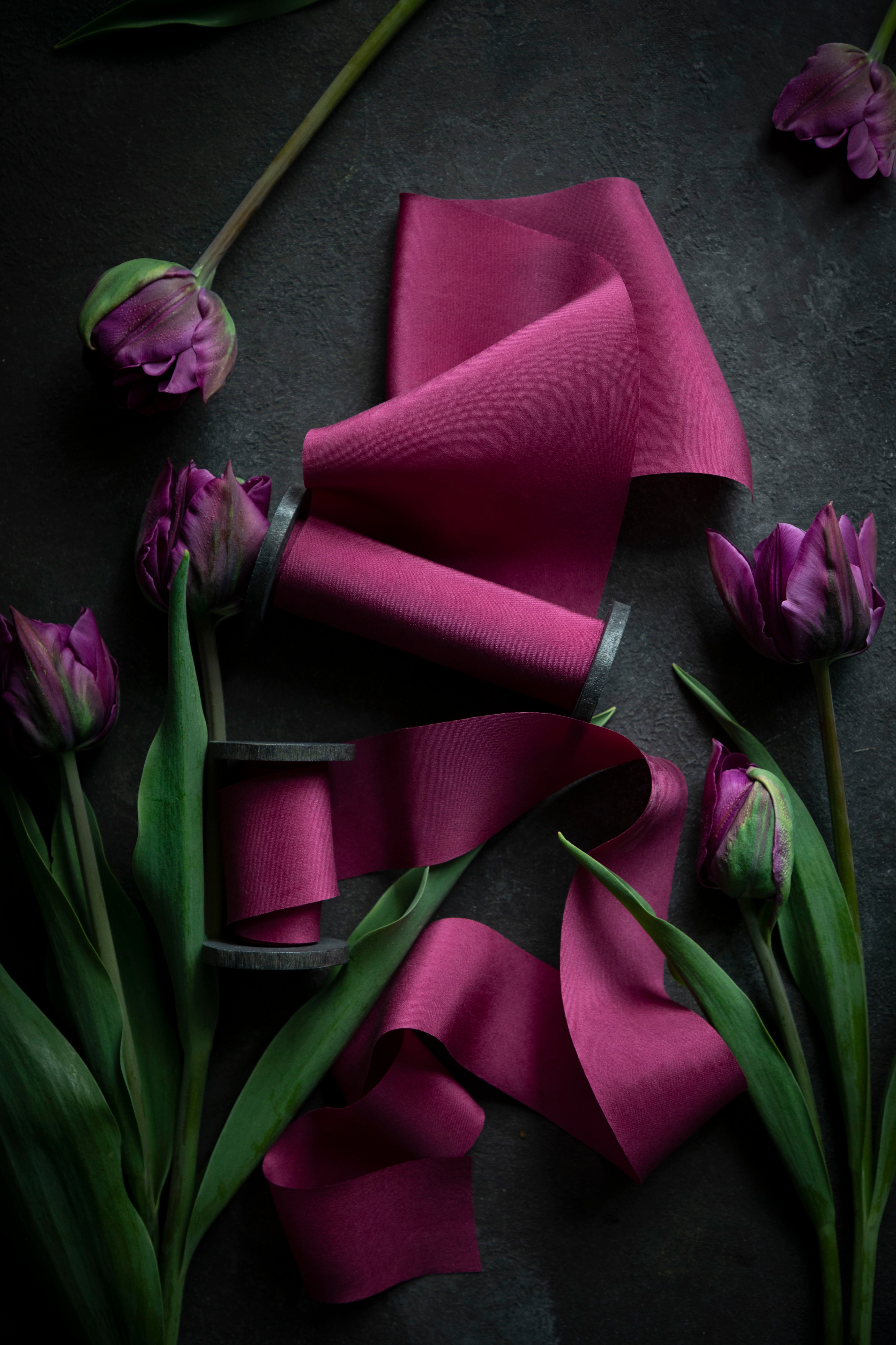 A series of vibrant pink silk ribbons arranged on a dark surface, surrounded by purple tulips, emphasizing the color and texture of the ribbons.