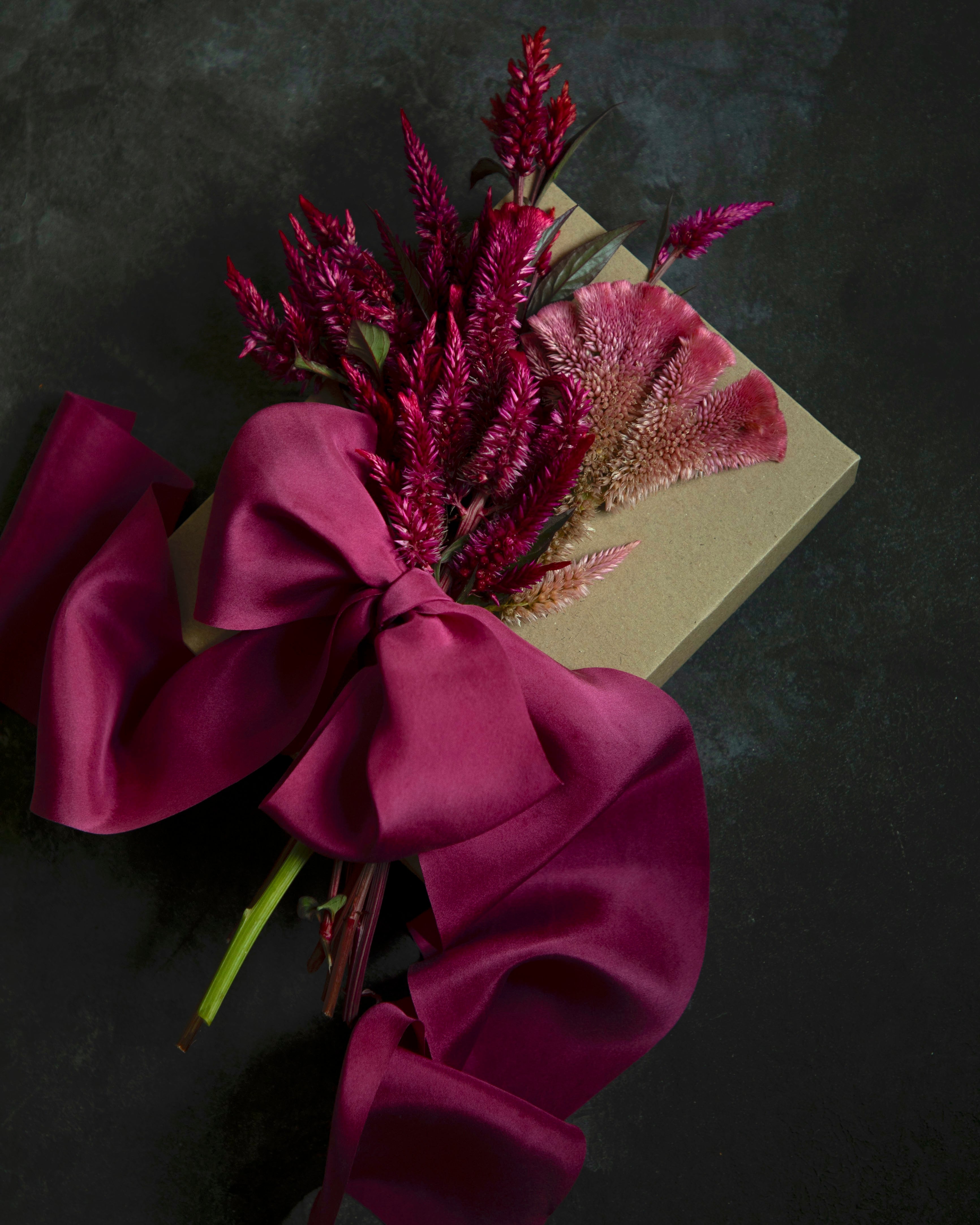 A dried floral arrangement featuring deep magenta dried flowers, a pinkish-purple feather grass flower, and a large magenta satin bow wrapped around a square kraft paper box, set against a dark textured background.