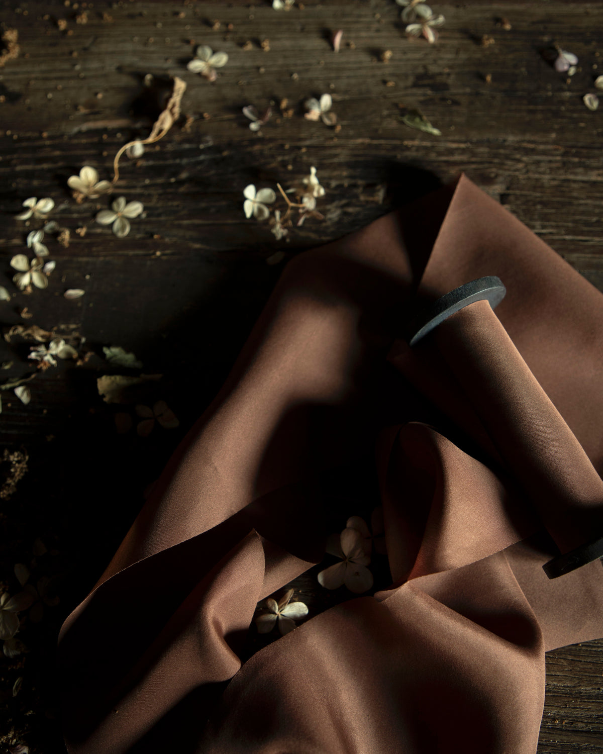 A close-up of draped brown ribbon on a wooden spool, arranged on a dark rustic wooden surface with scattered small white flowers.