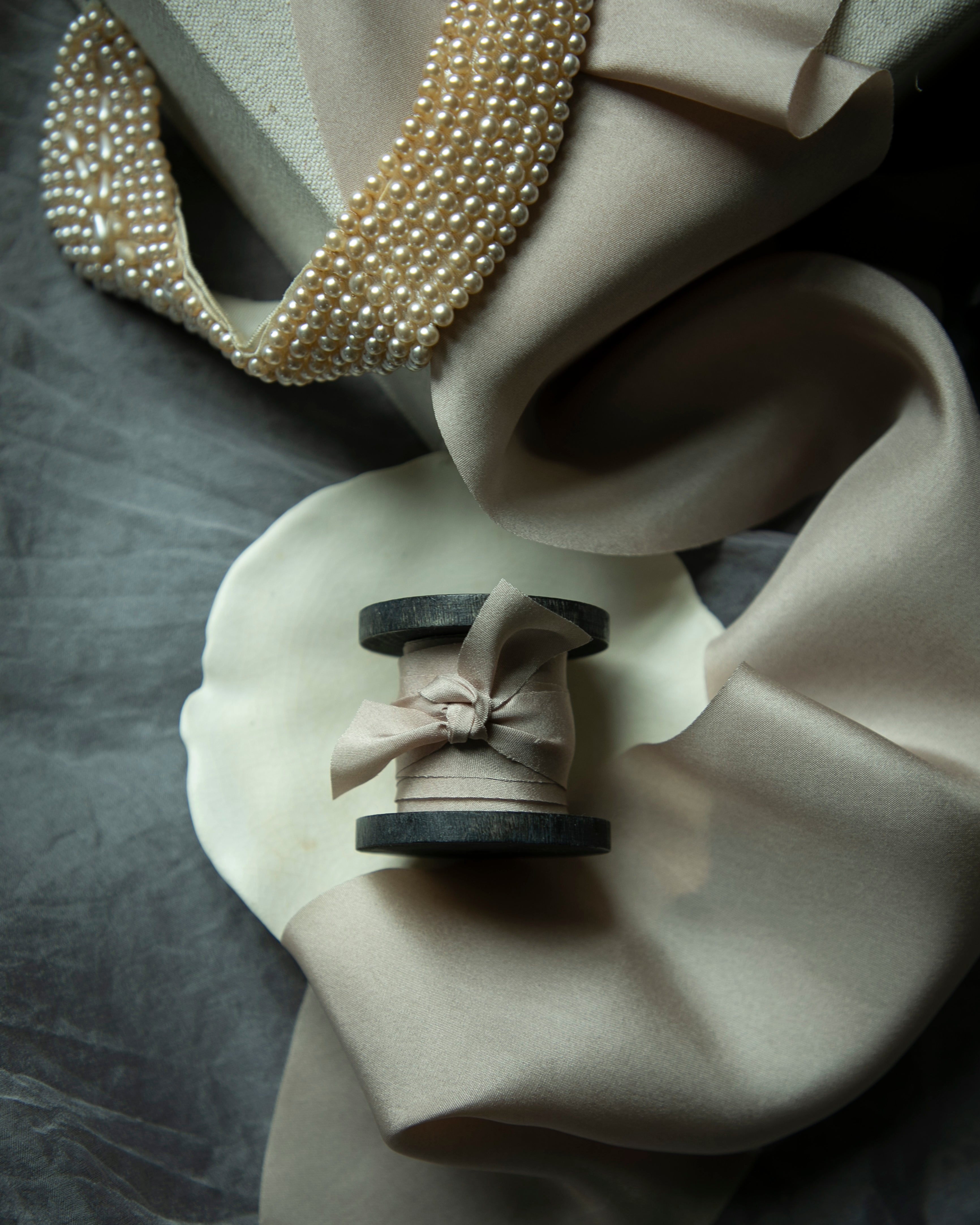 Pearl necklace draped over a beige fabric with a spool of thread and ribbon on a gray surface.