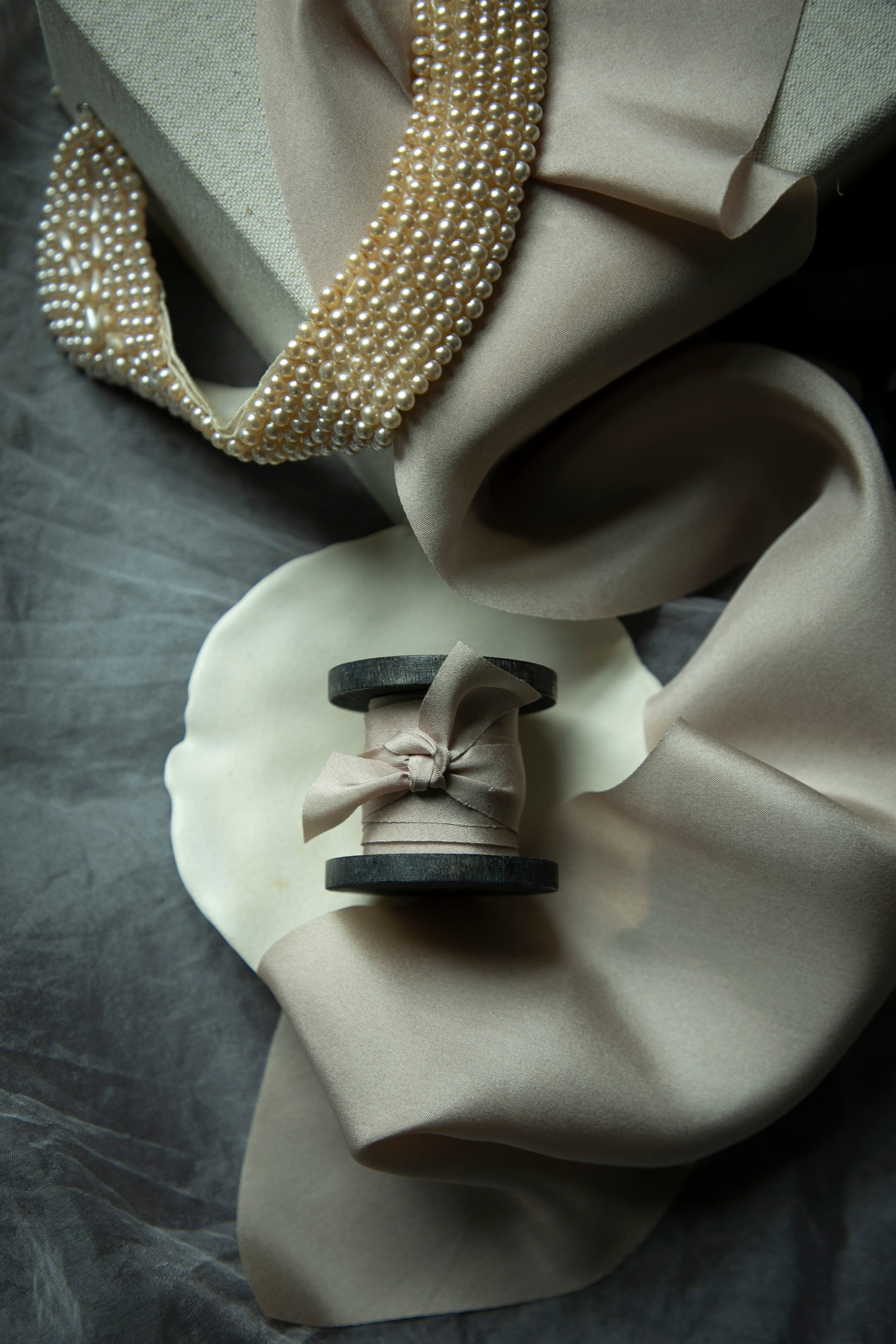Pearl necklace draped over a beige fabric with a spool of thread and ribbon on a gray surface.