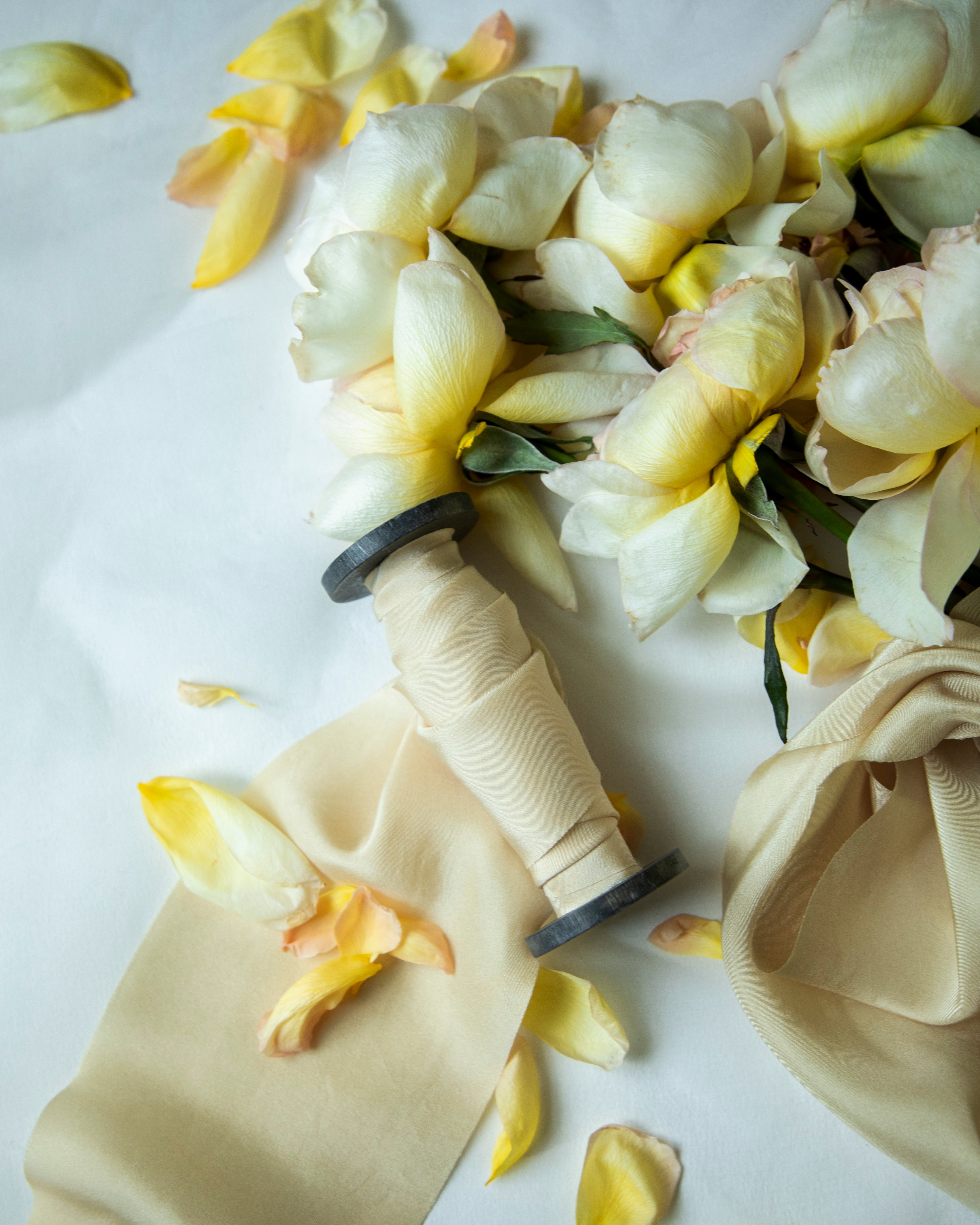 A bouquet of pale yellow and white flowers arranged on a white surface with a cream satin ribbon and scattered flower petals.