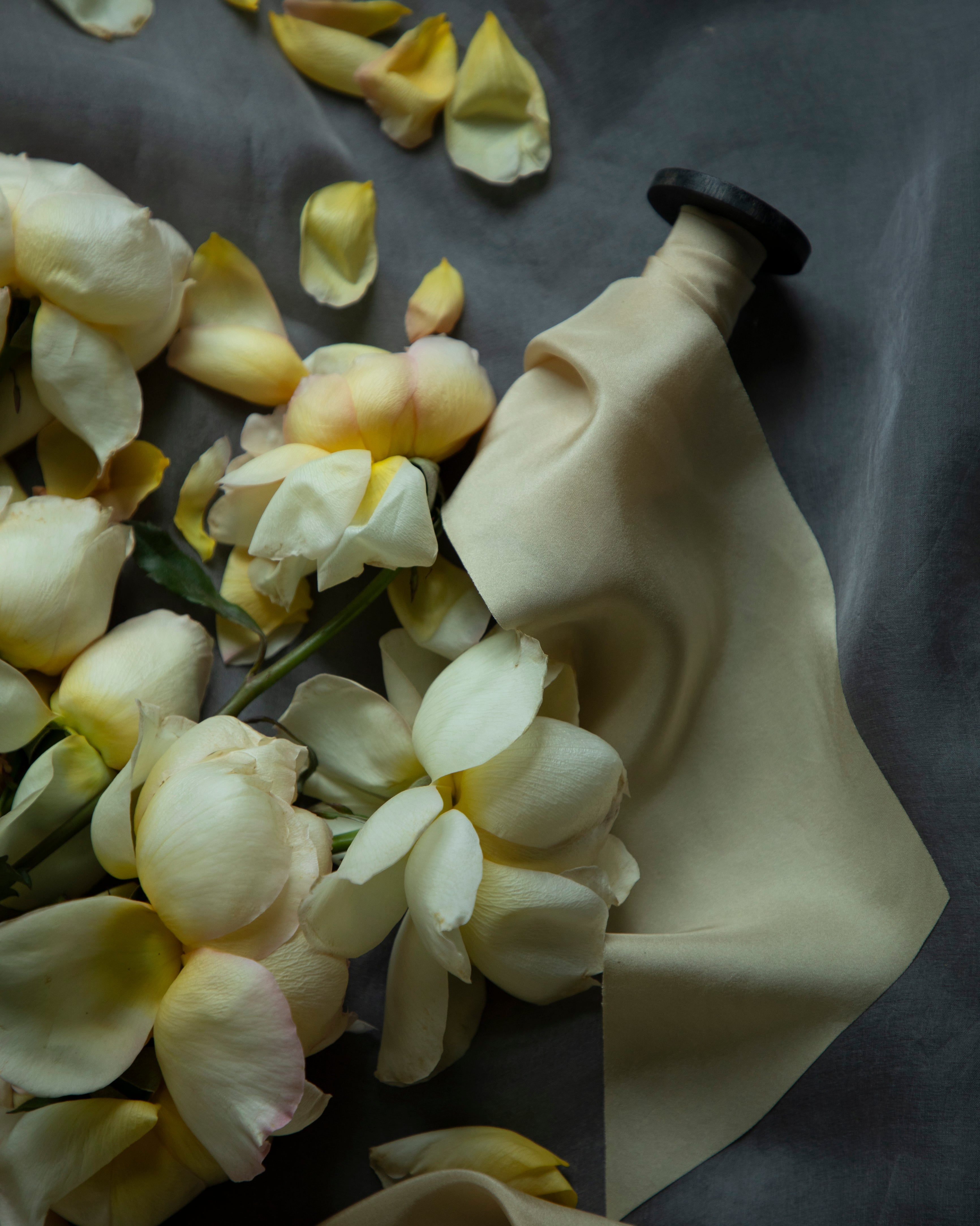 A antique gold silk ribbon spool with a black wood spool, shown with draped fabric and scattered pale yellow flower petals on a dark gray background.