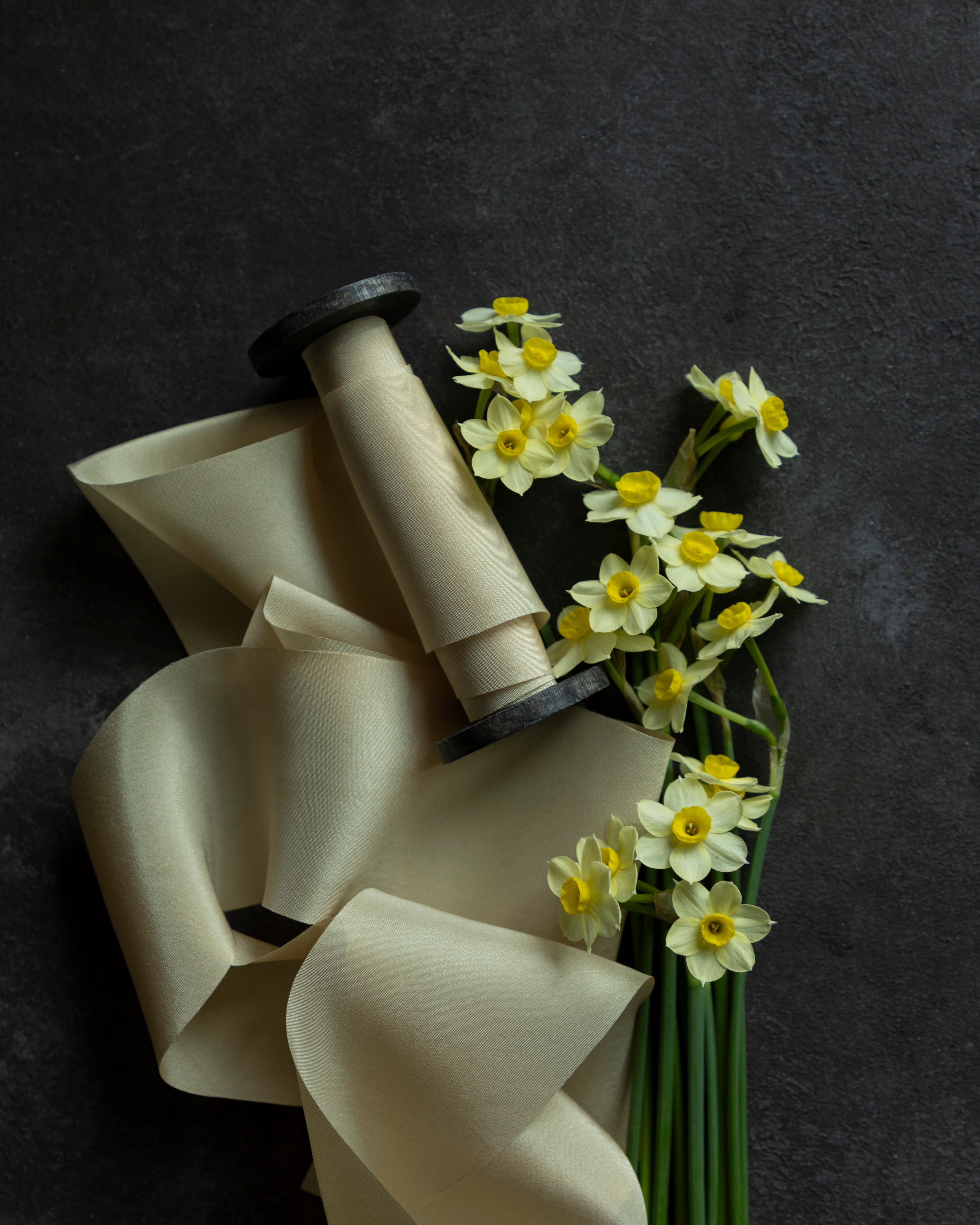 A spool of antique gold silk ribbon next to a bouquet of yellow flowers, arranged on a dark background.
