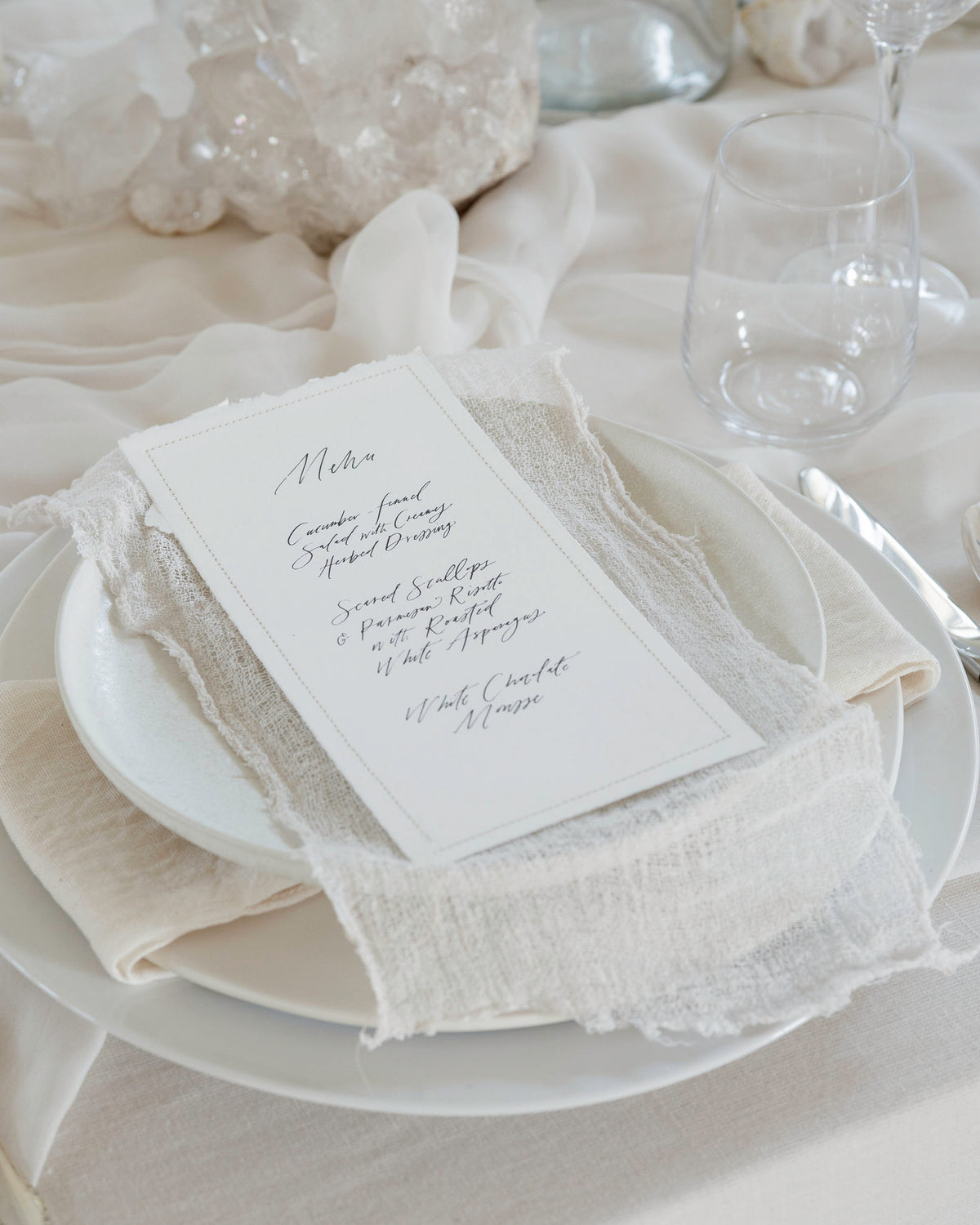A wedding place card with elegant black script lettering, styled on a white plate with a cream textured linen napkin and matching tablecloth.