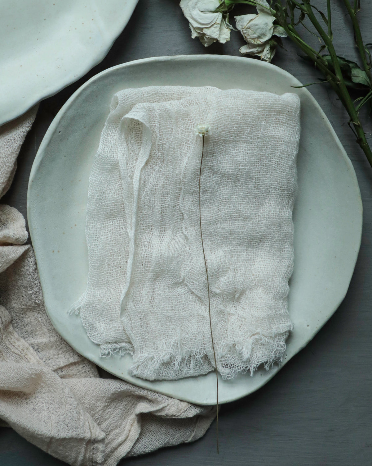 A folded off-white linen cloth napkin with frayed edges rests on a round ceramic plate, styled with dried white roses and neutral fabric on a dark gray tabletop.