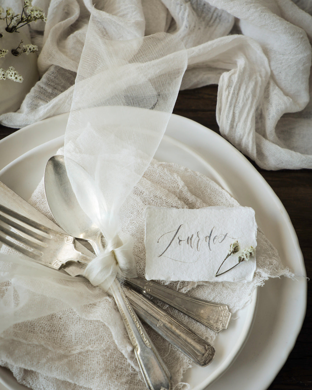 A styled place setting on a white plate with silver-toned flatware, a small white napkin, and a paper place card reading 'Journée,' wrapped in sheer white fabric against a dark wood background with draped white cloth.