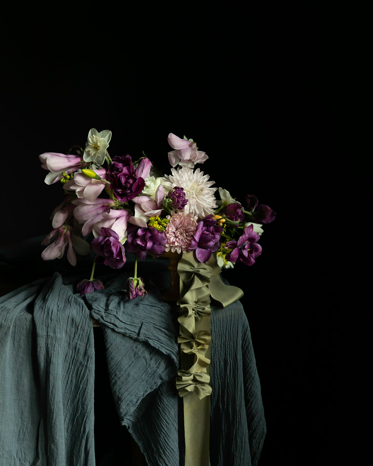 A floral arrangement with purple, pink, and white flowers draped over dark teal and olive-green textured fabric against a solid black background.