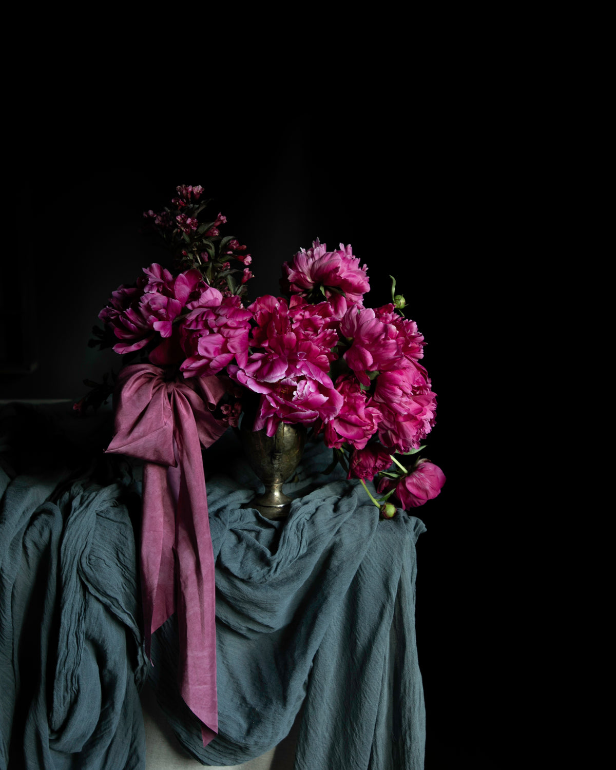 A bouquet of vibrant magenta peonies in a small metallic vase, draped with a dusty blue-gray silk scarf against a solid black background.