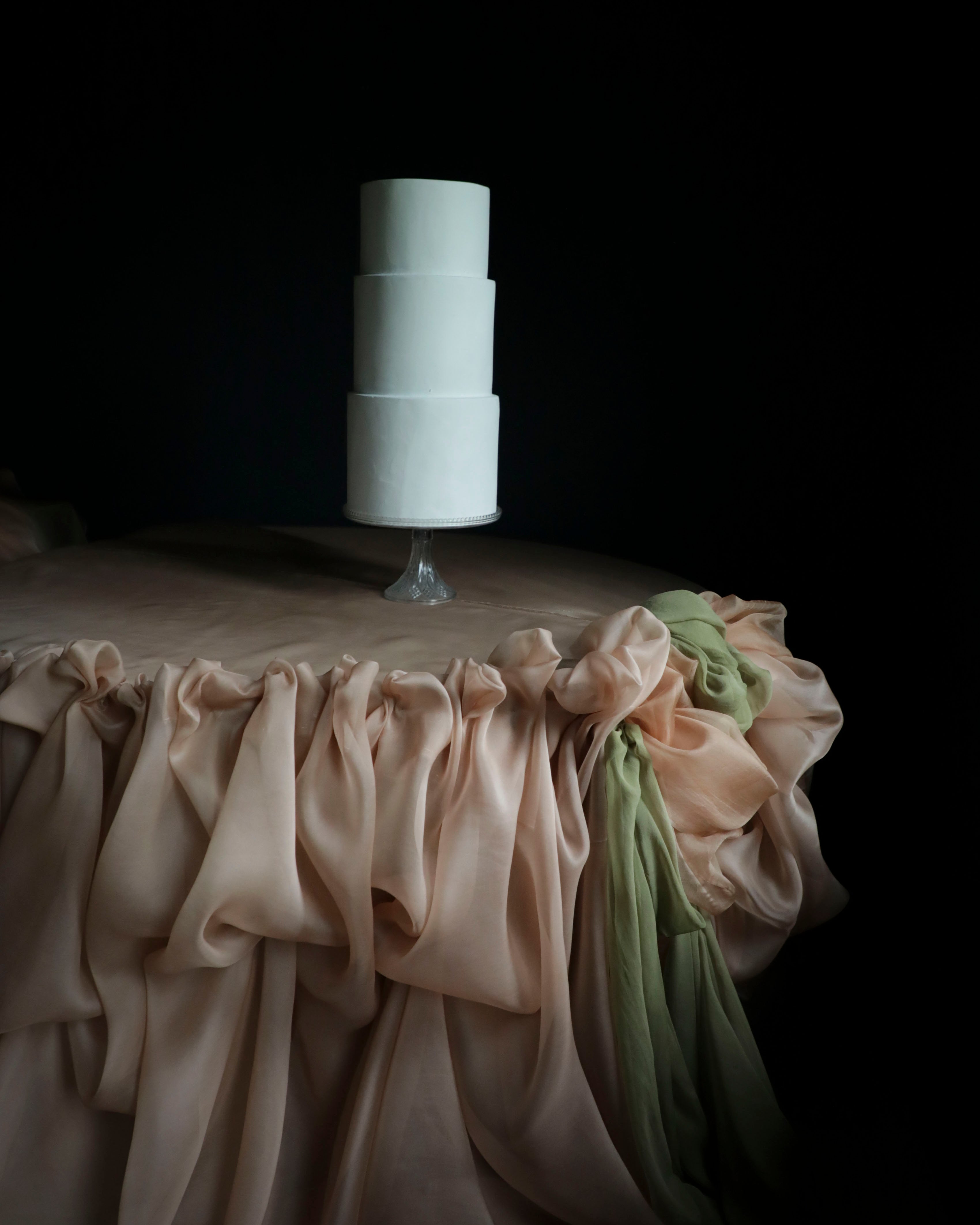 A three-tier white cake on a clear stand, set against a dark background with a draped champagne tablecloth and a green fabric accent.
