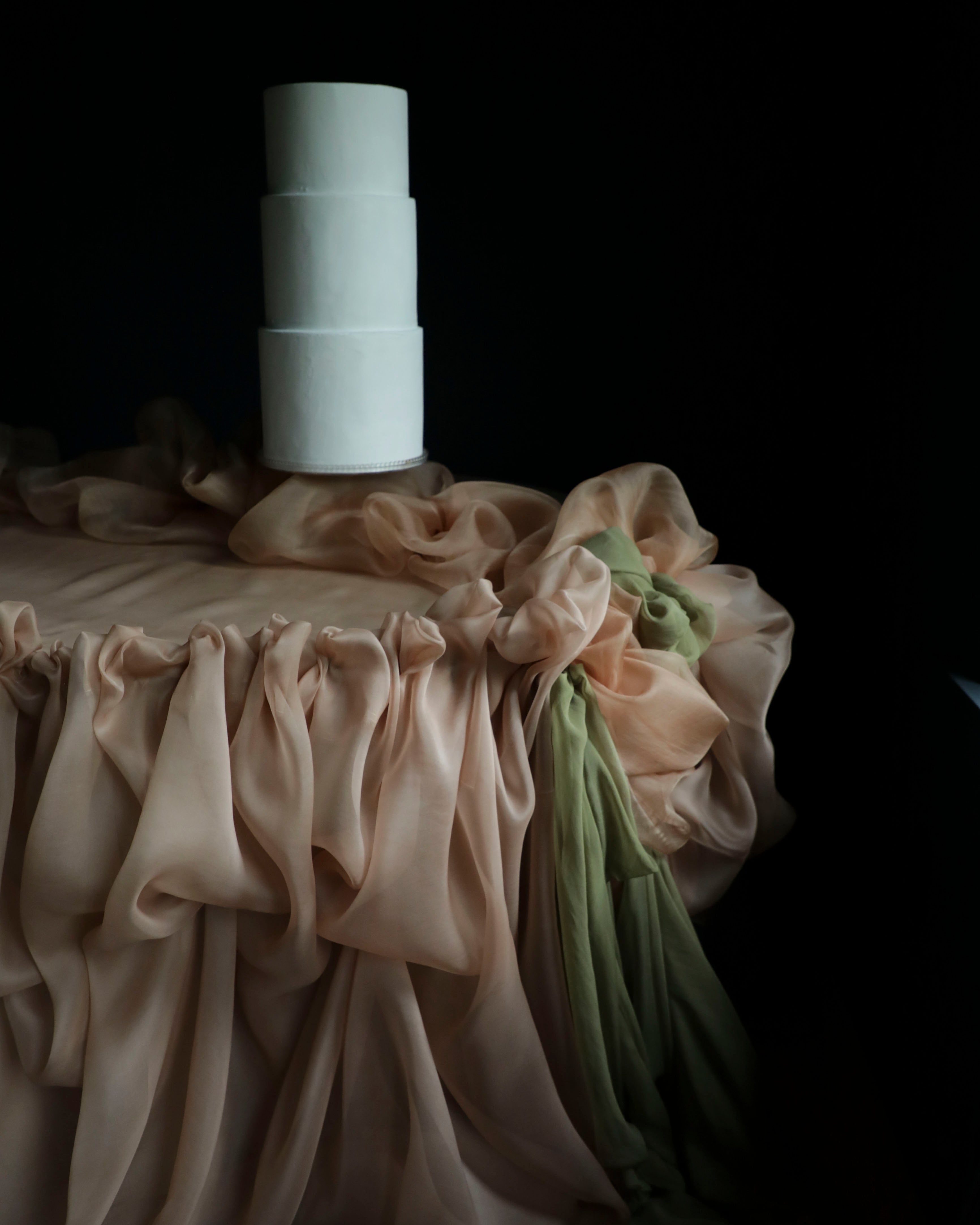 A white wedding cake on a draped surface covered in soft blush silk fabric with a green section, set against a very dark background.
