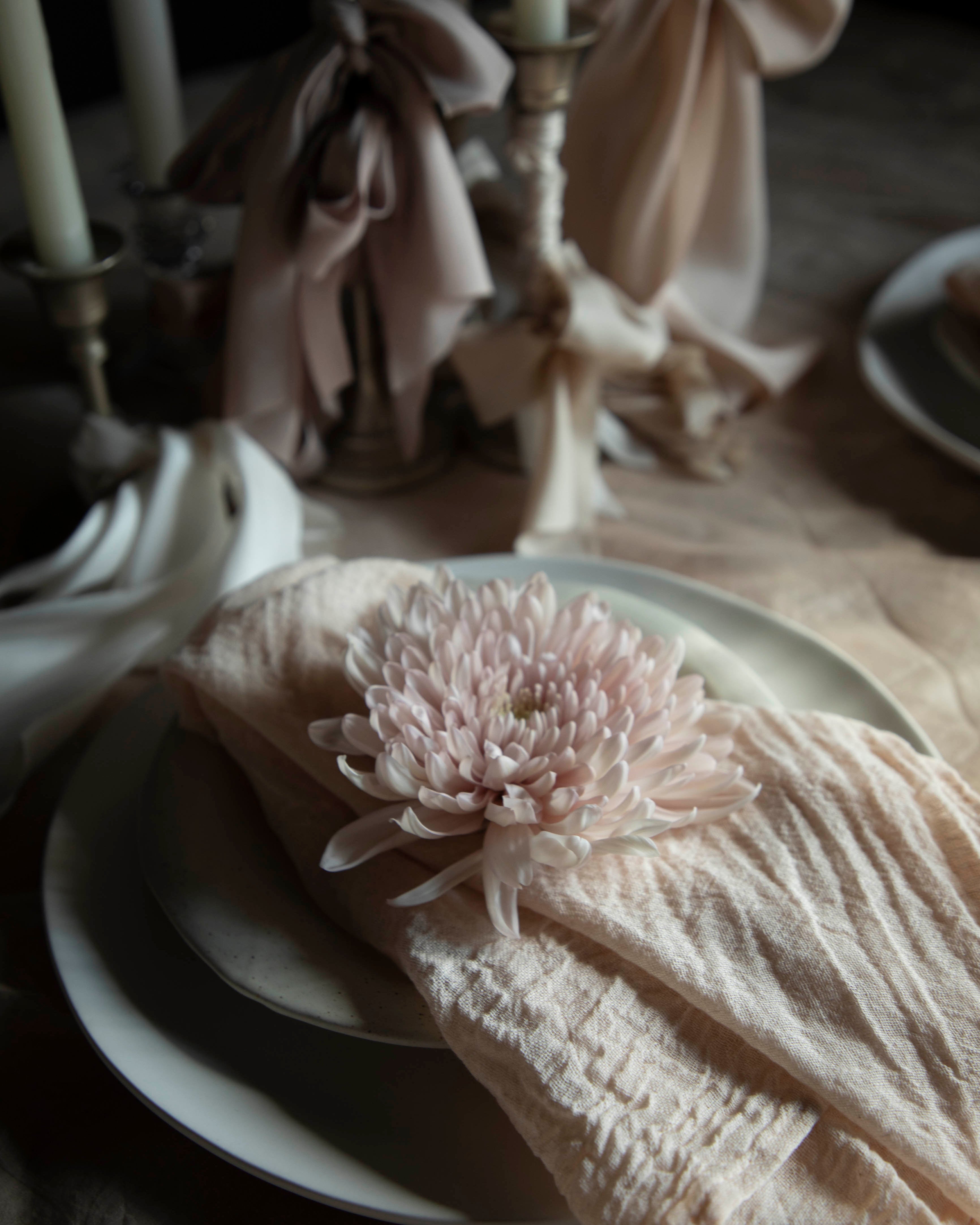 Neatly folded beige napkin with a decorative flower on a white plate, set against a blurred background of candles and bows.
