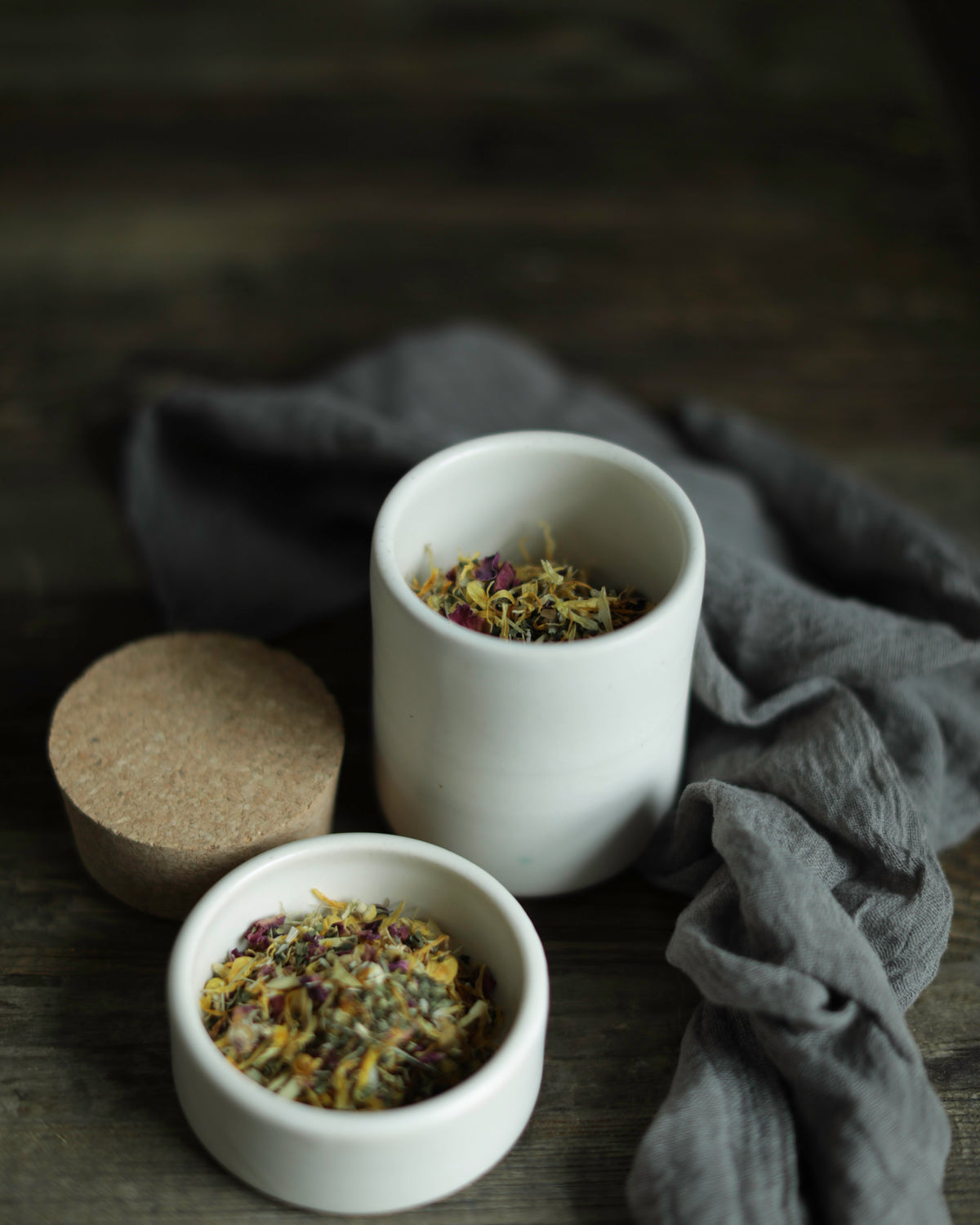 Two small white ceramic cups filled with loose herbal tea, placed on a dark wooden surface with a gray cloth and a cork lid nearby.