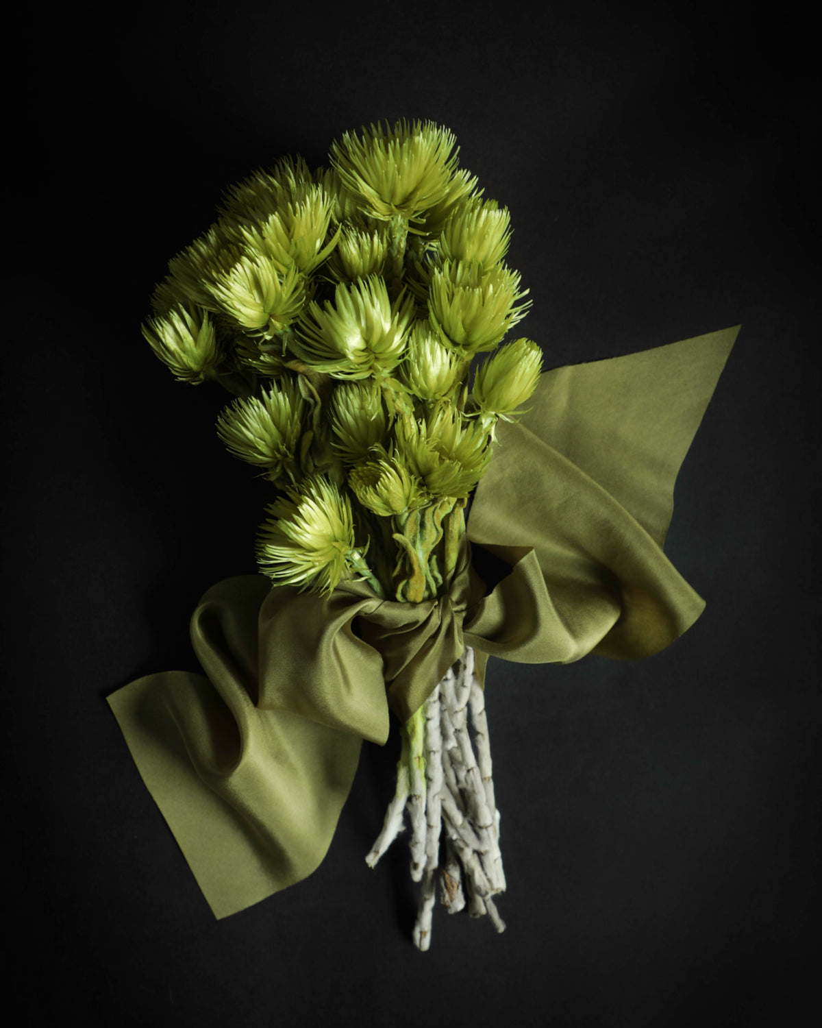 A bouquet of light green dried flowers wrapped in olive green paper and tied with a matching ribbon, set against a solid black background.