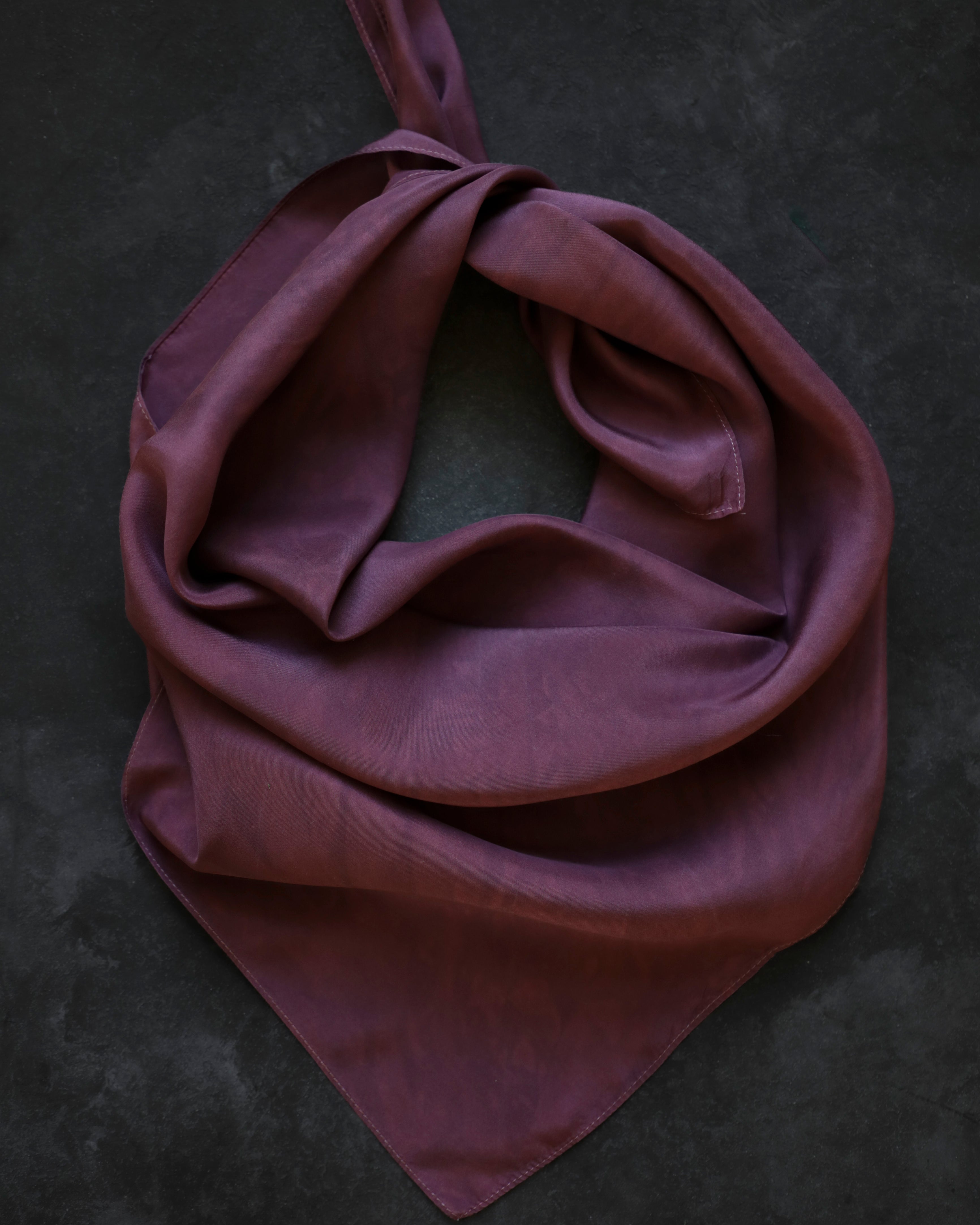 A mauve-purple silk scarf tied in a loose knot, displayed on a dark textured background.