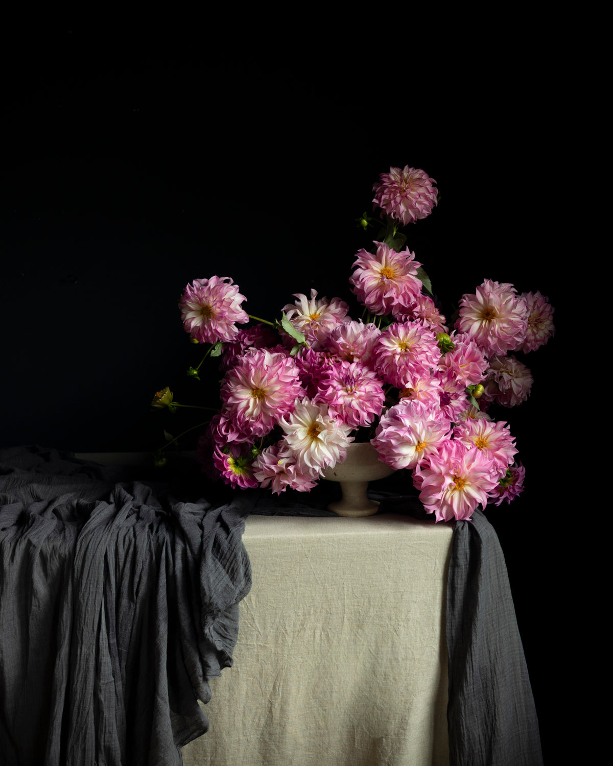 A bouquet of pink and white dahlia flowers arranged in a ceramic vase on a draped table, set against a dark black background with a light-colored linen fabric covering the table.