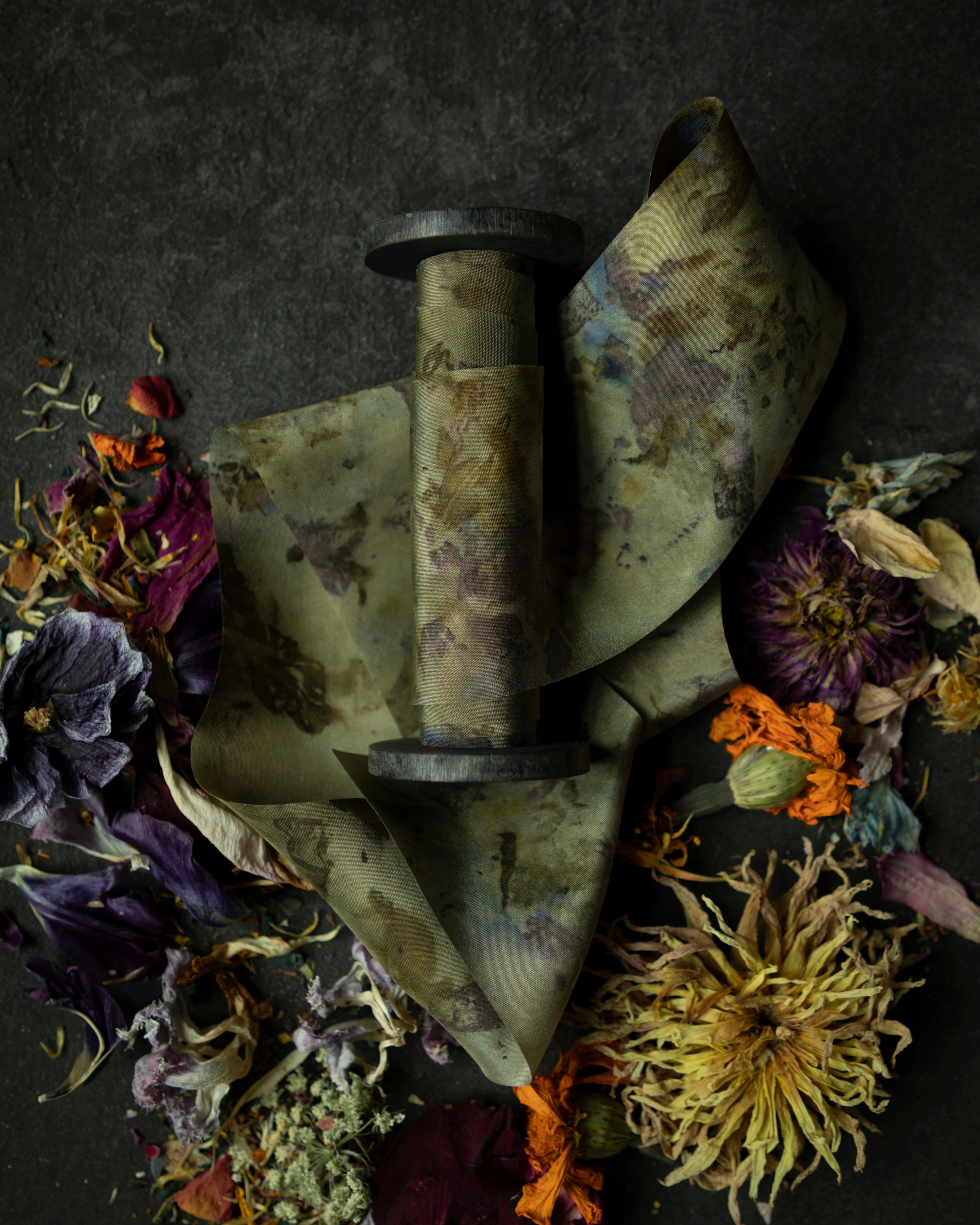 Limited-edition eco-print silk ribbon displayed on a wood spool surrounded by an assortment of dried flowers