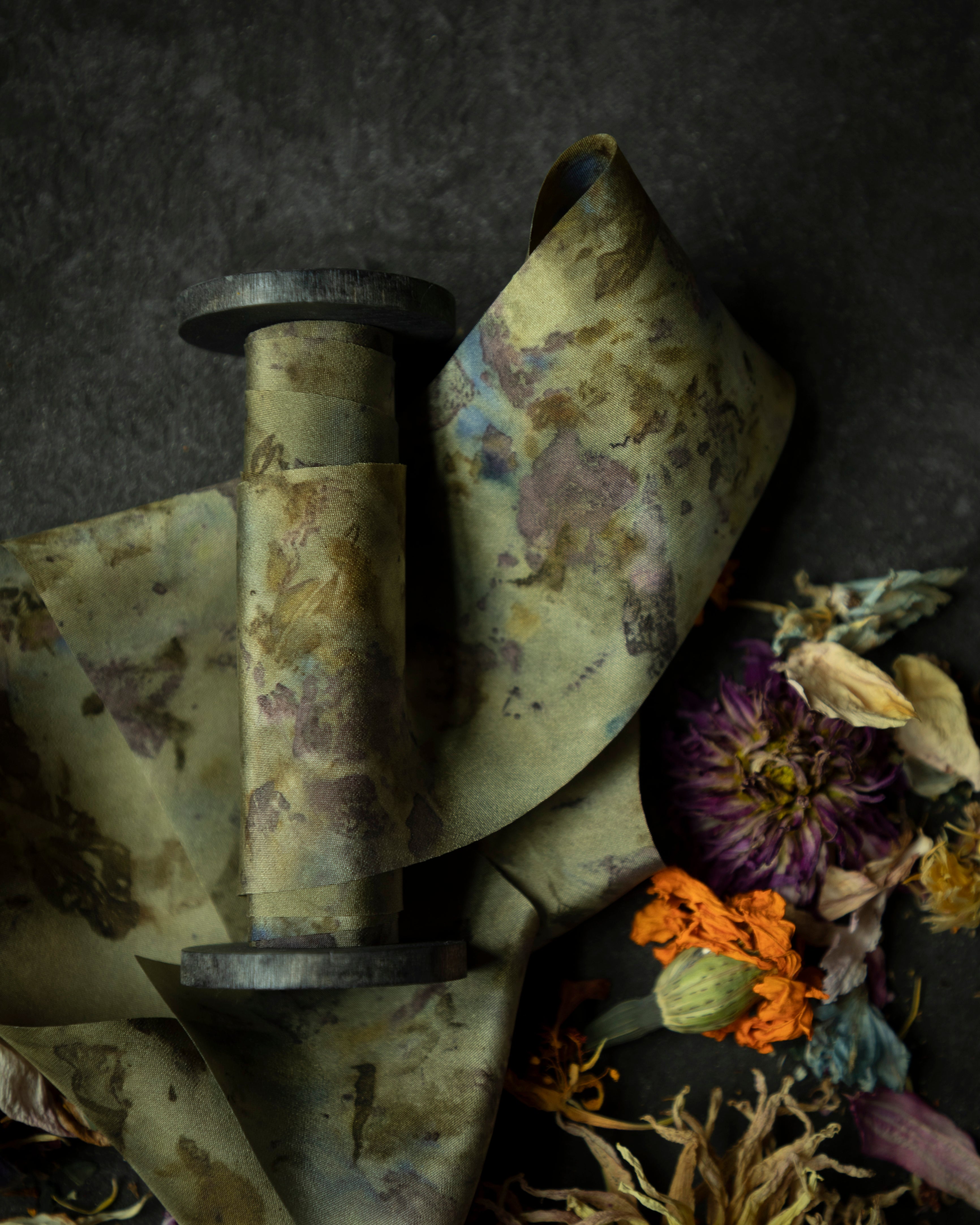 Limited-edition eco-print silk ribbon displayed on a wood spool surrounded by an assortment of dried flowers