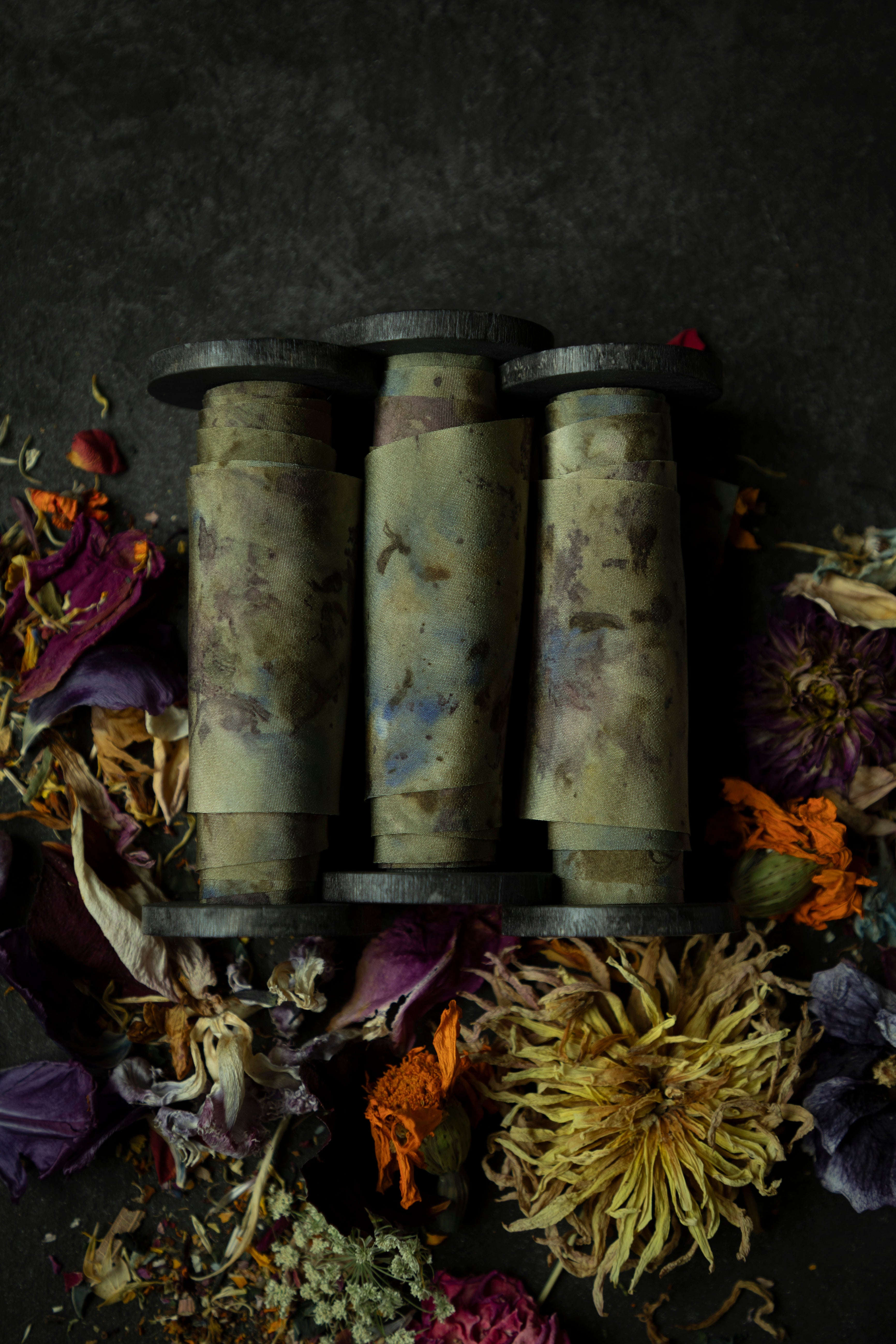 Limited-edition eco-print silk ribbon displayed on a wood spool surrounded by an assortment of dried flowers