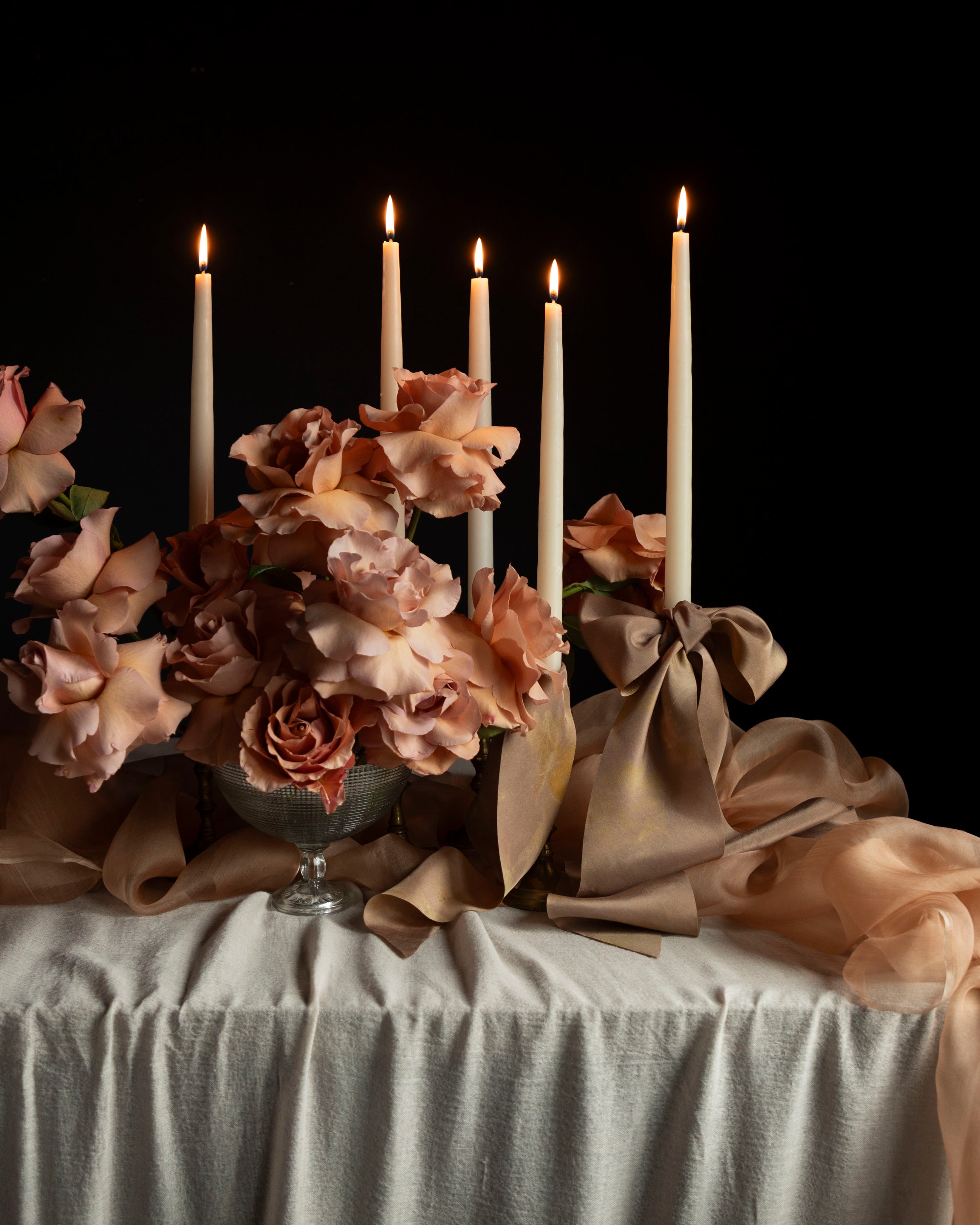 Decorative floral arrangement with coral roses and candles on a handmade tablecloth against a black background
