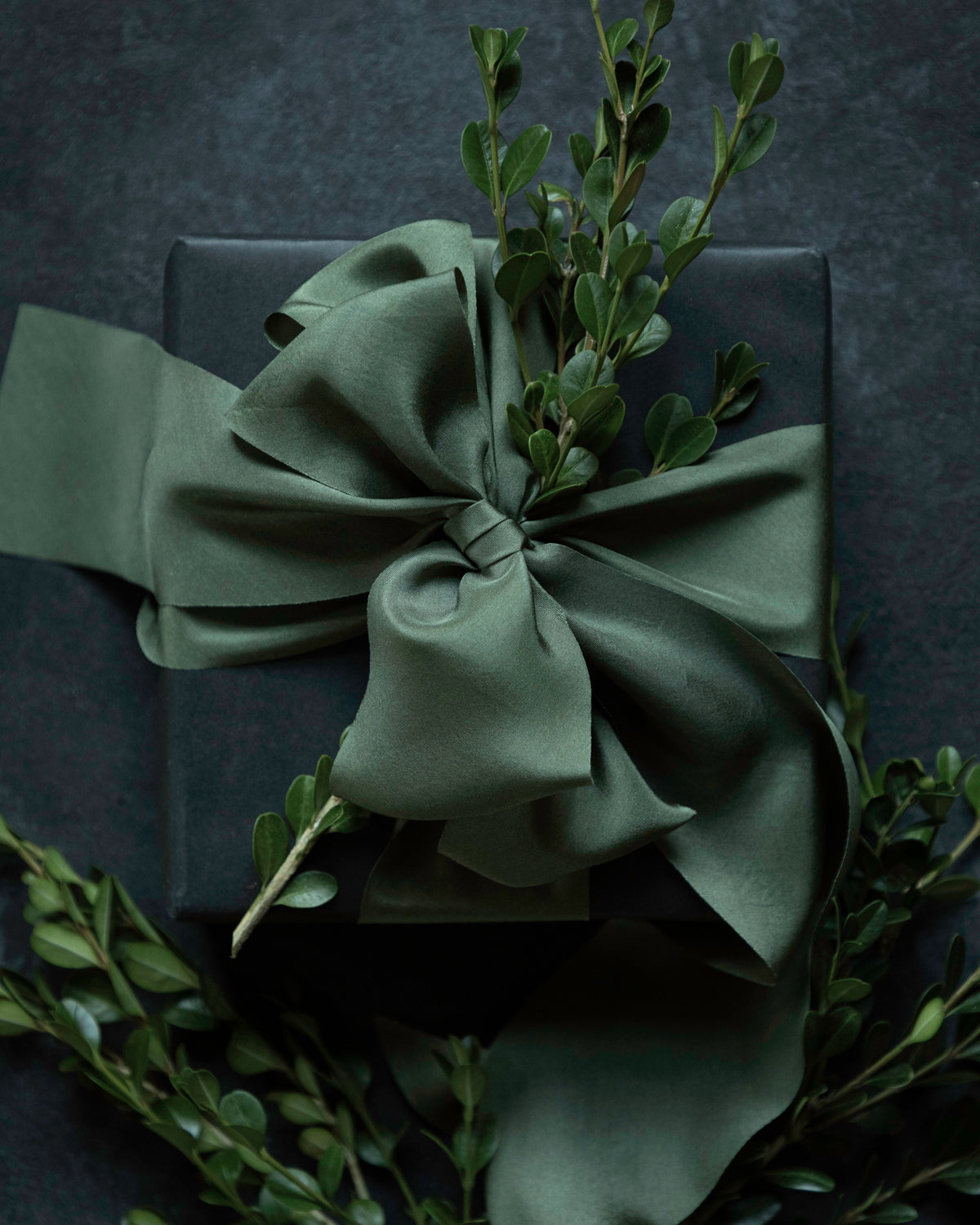 An olive green satin ribbon tied in a large bow around a dark gift box, decorated with green foliage, set against a dark textured background. Luxury gift Wrapping