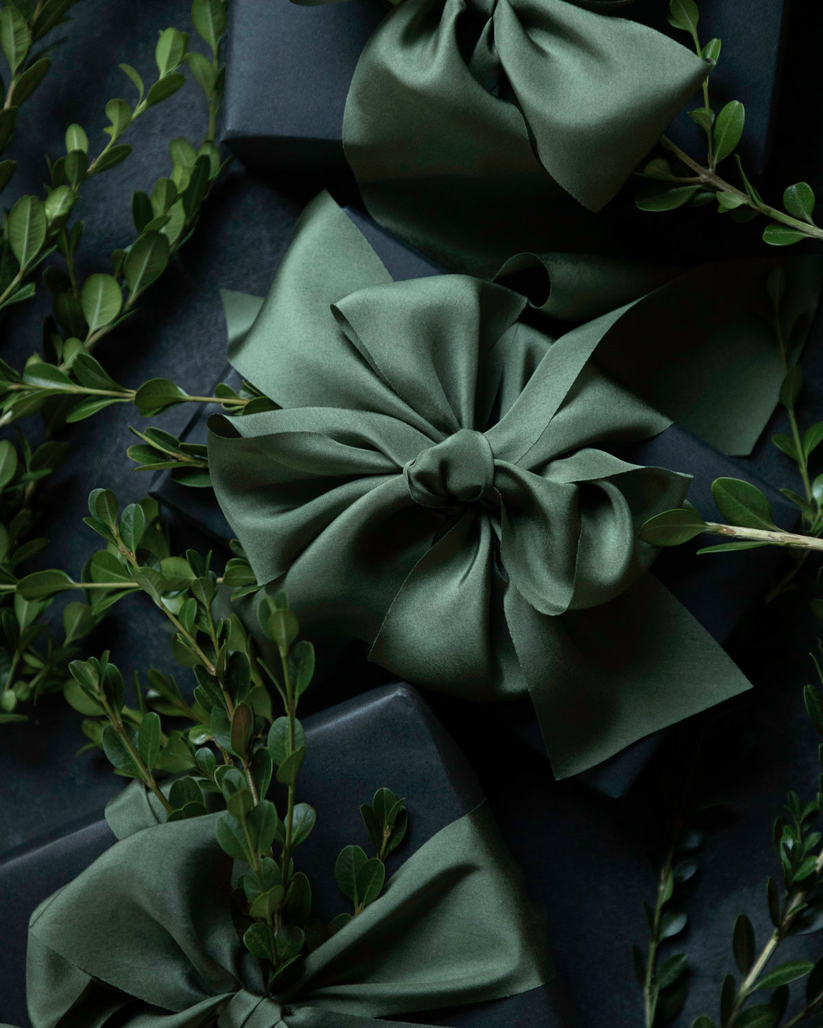 Three dark green satin gift bows tied around dark navy wrapped boxes, styled with green foliage on a dark background.