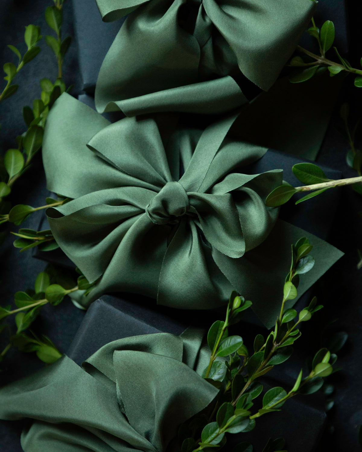Three large satin bows in a dark sage green color, tied around dark wrapped gift boxes and accented with fresh green leaves, set against a dark background.