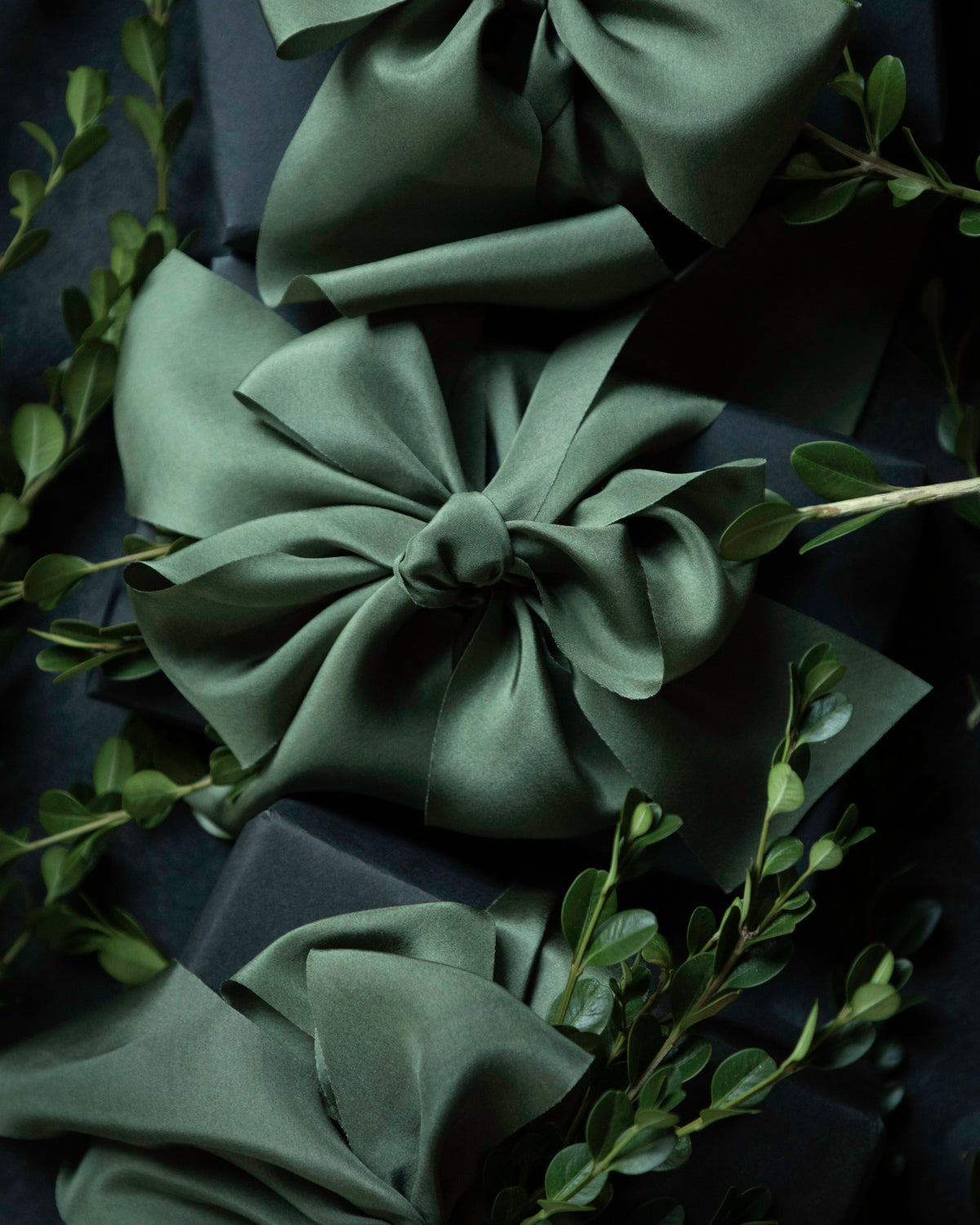 Three large satin gift bows in a muted sage green color, tied around dark wrapped boxes and accented with delicate green foliage. Gift wrapping inspiration