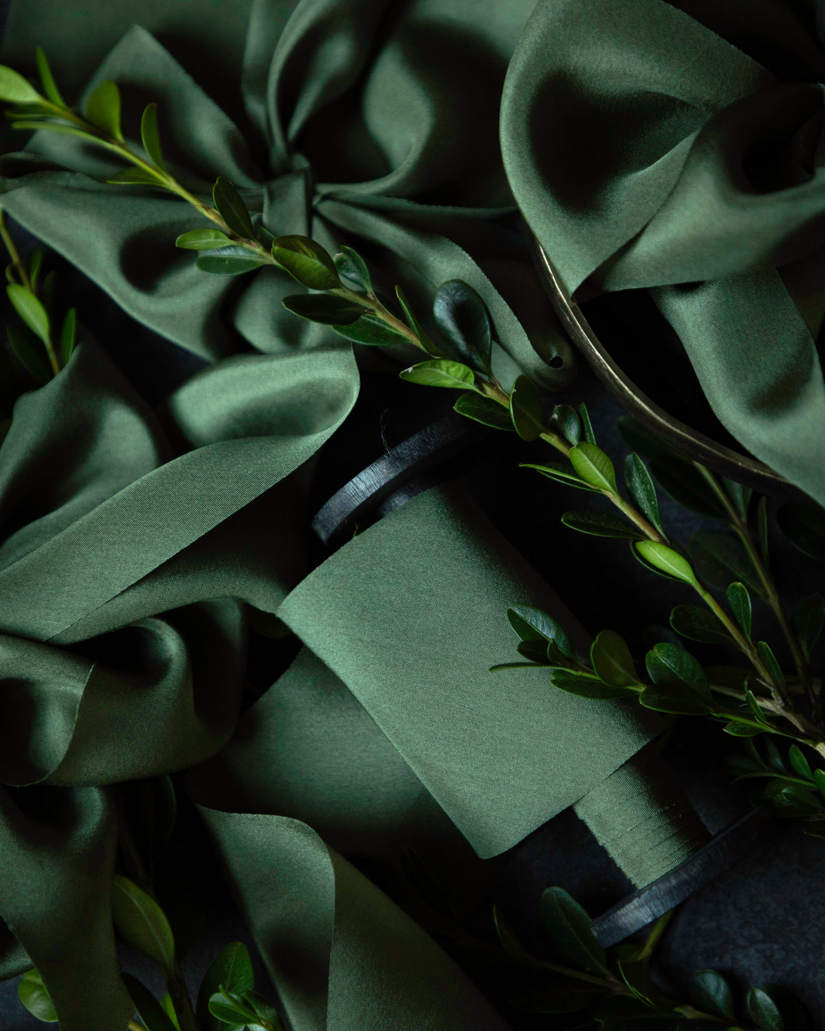Dark green satin ribbon and matching fabric napkins styled with fresh green leaves on a dark background.