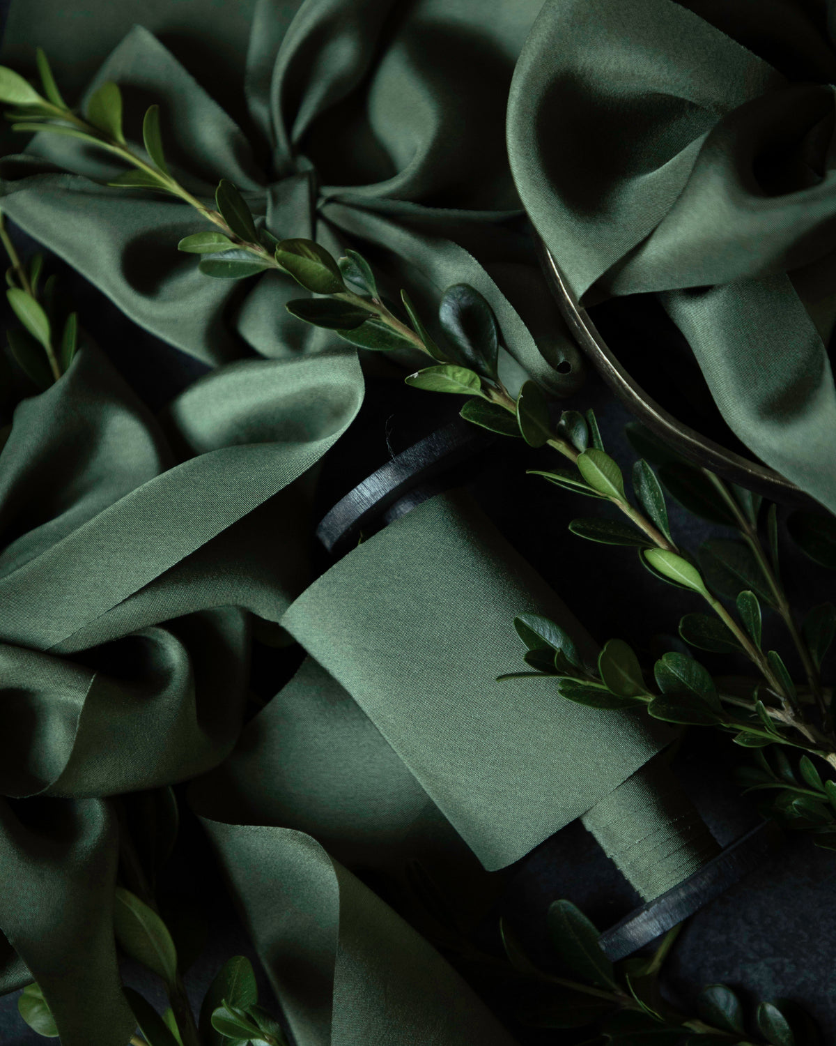 Dark green satin ribbon arranged with small green boxwood leaves on a dark background.