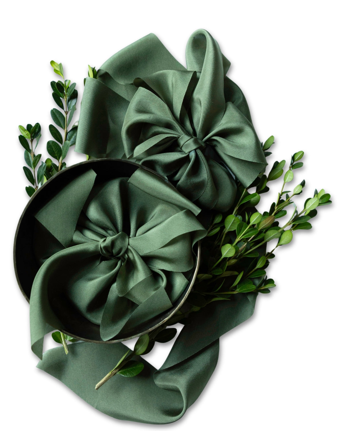 A dark green satin bow and matching ribbon arranged in a decorative flat lay with small green leaves on a white background.