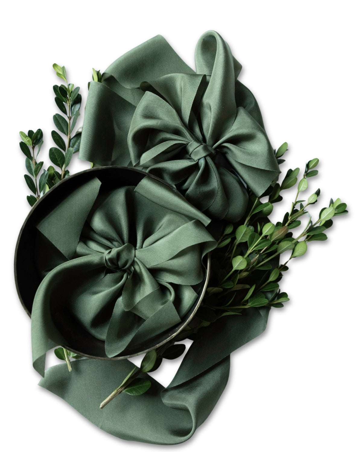 A dark sage green satin ribbon arranged in two large bows, styled with green boxwood branches on a plain white background.