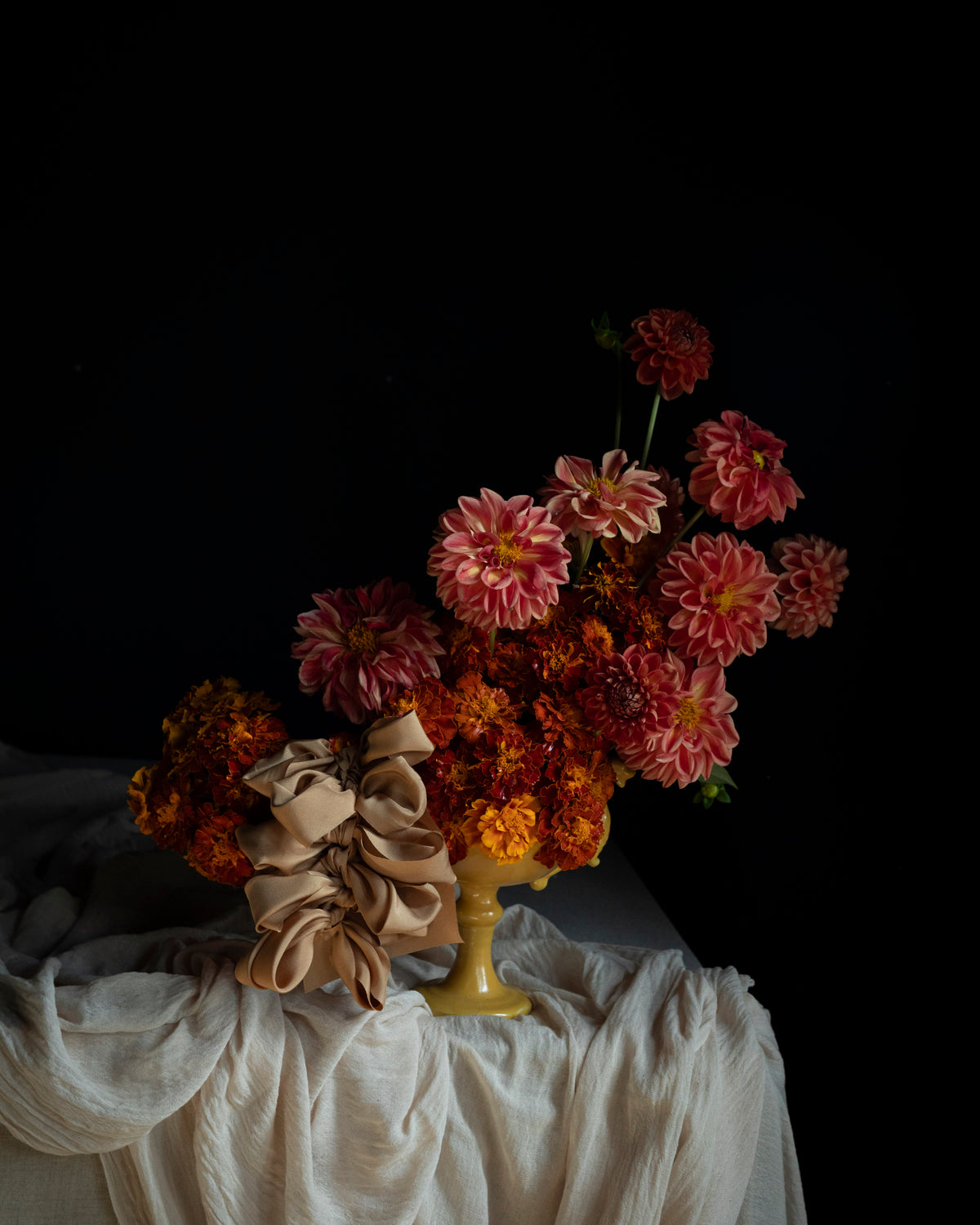 A still life floral arrangement featuring pink and orange dahlias with a beige ribbon bow, displayed in a gold pedestal vase draped with white fabric against a solid black background.