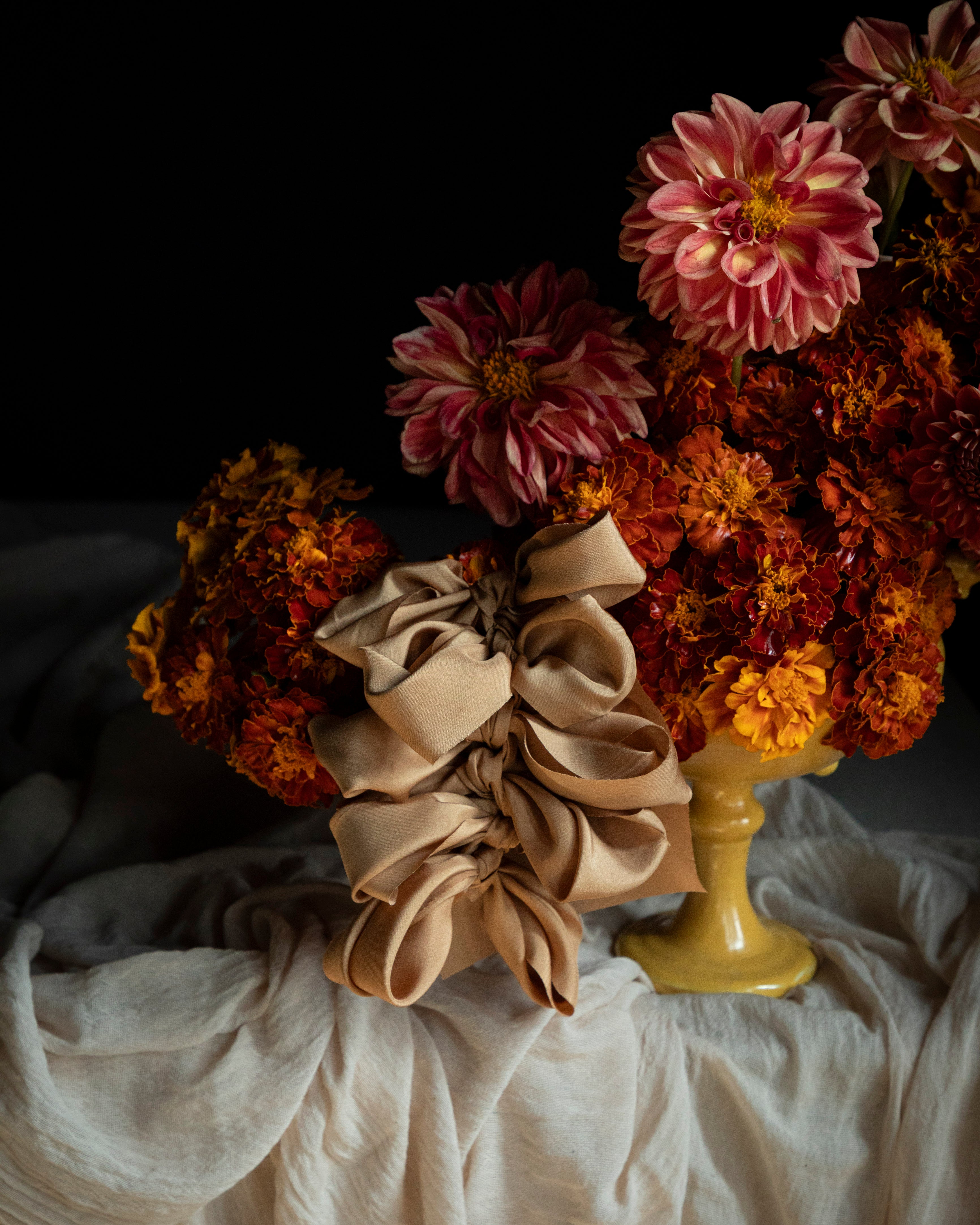 A styled still life featuring a large cinnamon-coral silk ribbon bow in the foreground, with a bouquet of orange, dahlia and marigold flowers in a yellow vase. On a draped white fabric against a black background.