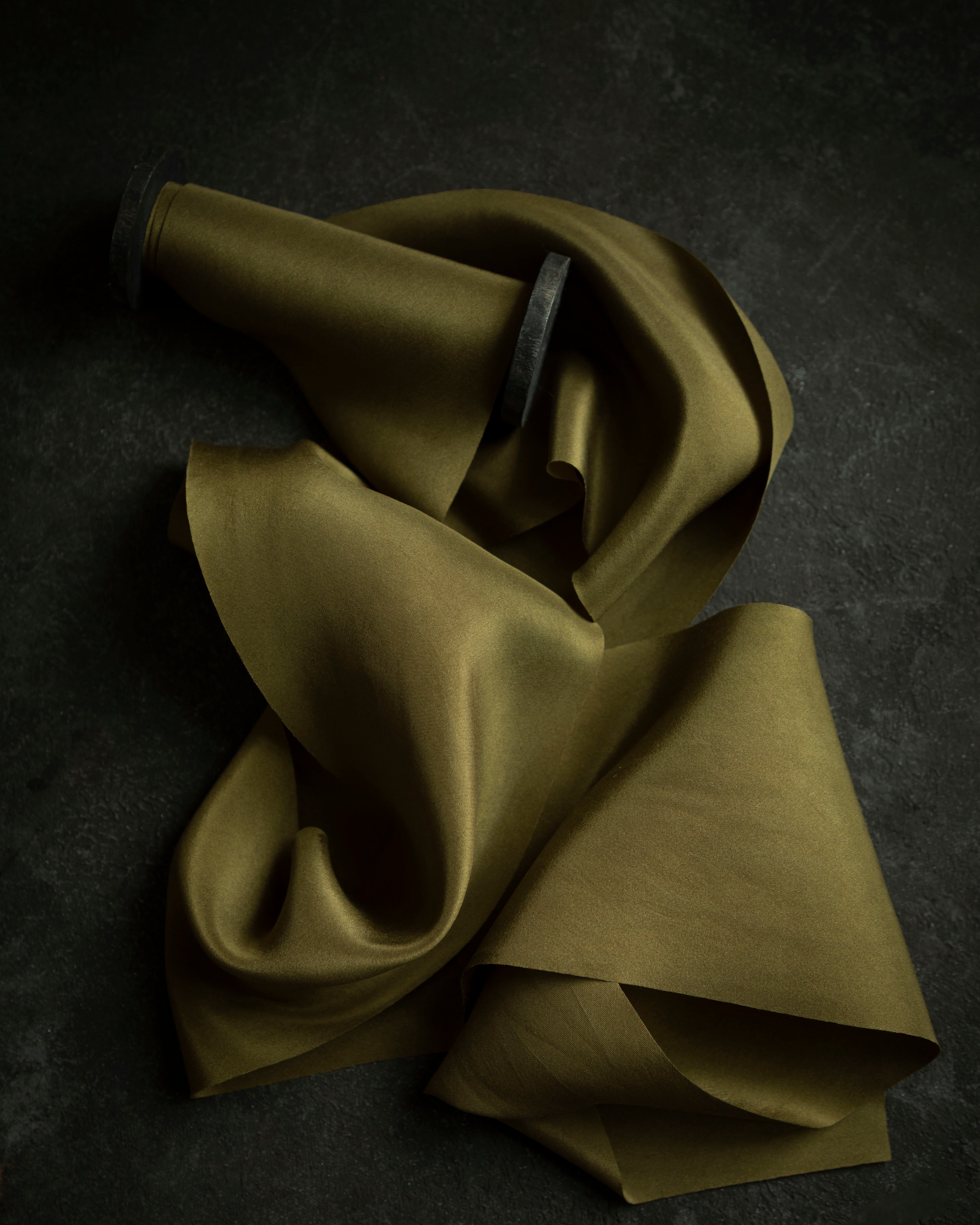 A Forest green silk ribbon with subtle olive-yellow undertones, laying on a dark surface, demonstrating its elegant and sophisticated texture.