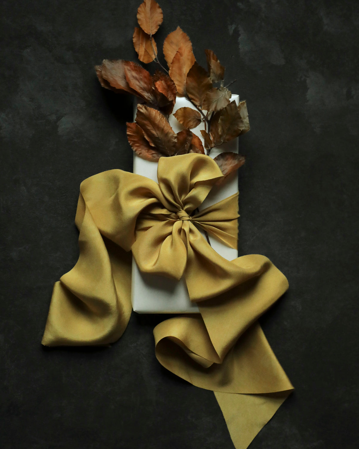 A white rectangular box wrapped with a large satin gold ribbon and bow, topped with dried brown leaves, arranged on a dark textured background.
