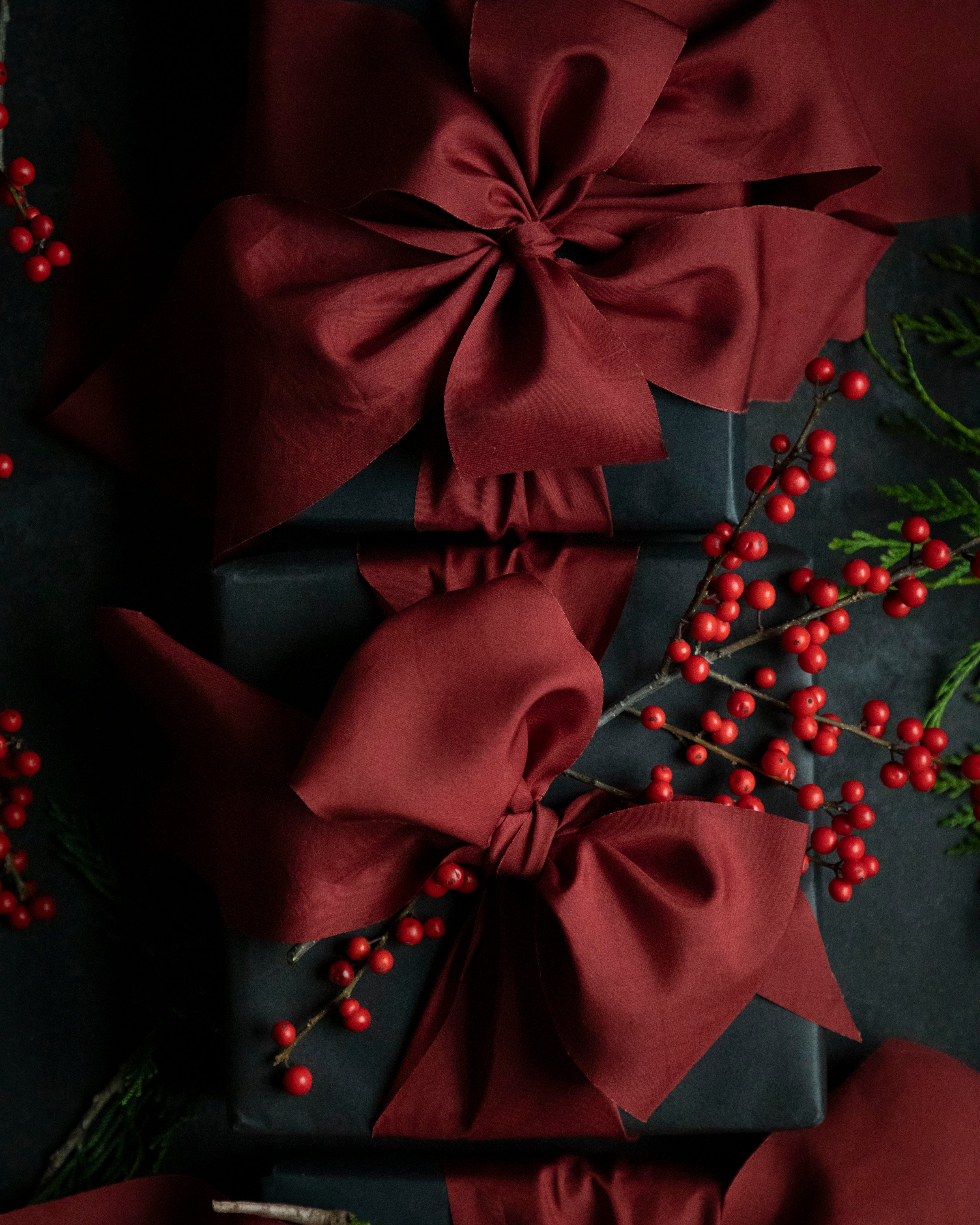 Gift wrapped in dark paper with a large red silk ribbon bow, surrounded by red berries and greenery on a dark background.