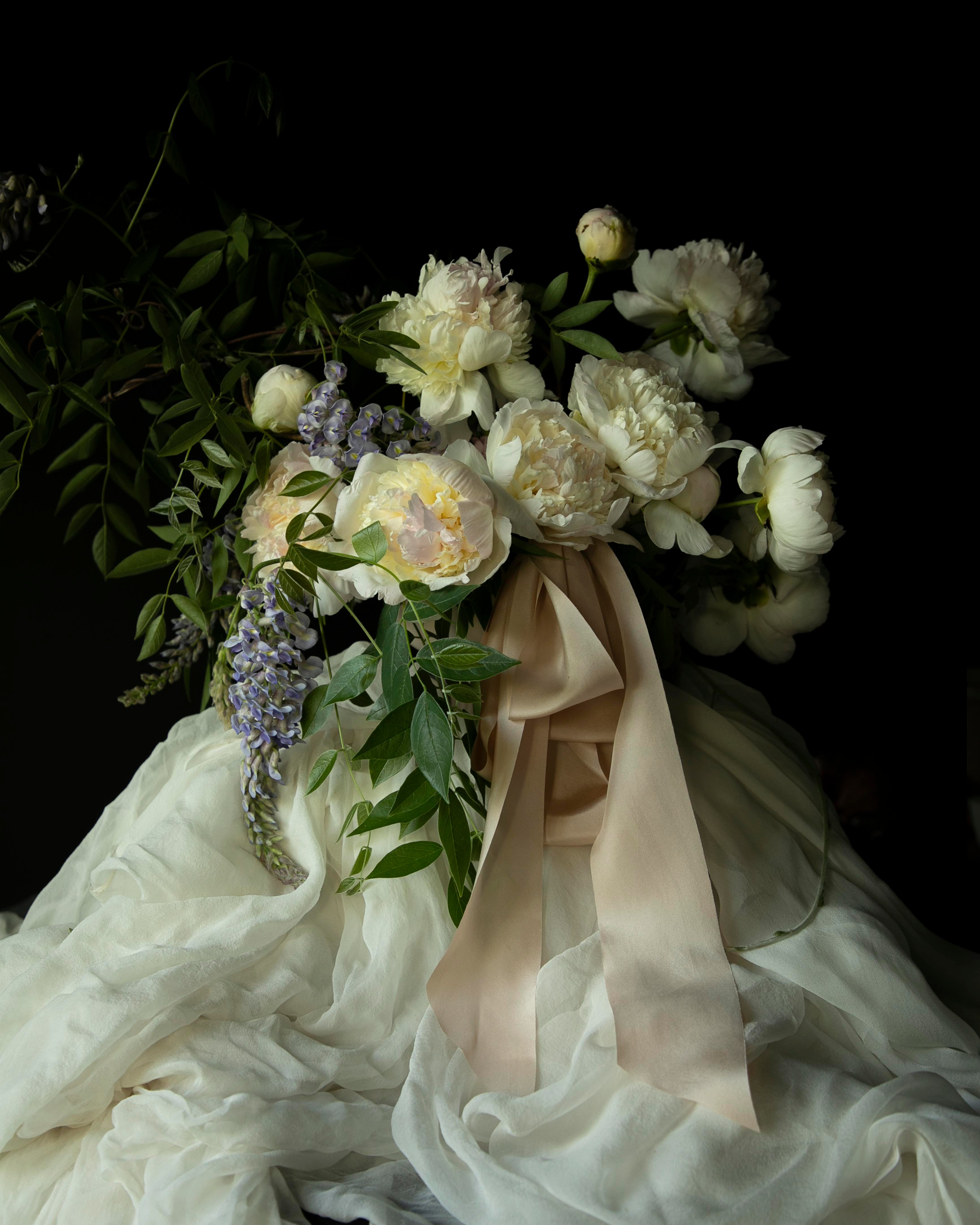 Floral arrangement with white flowers and green leaves draped over a white fabric against a dark background. highlighting the best wedding silk ribbon in blush color.