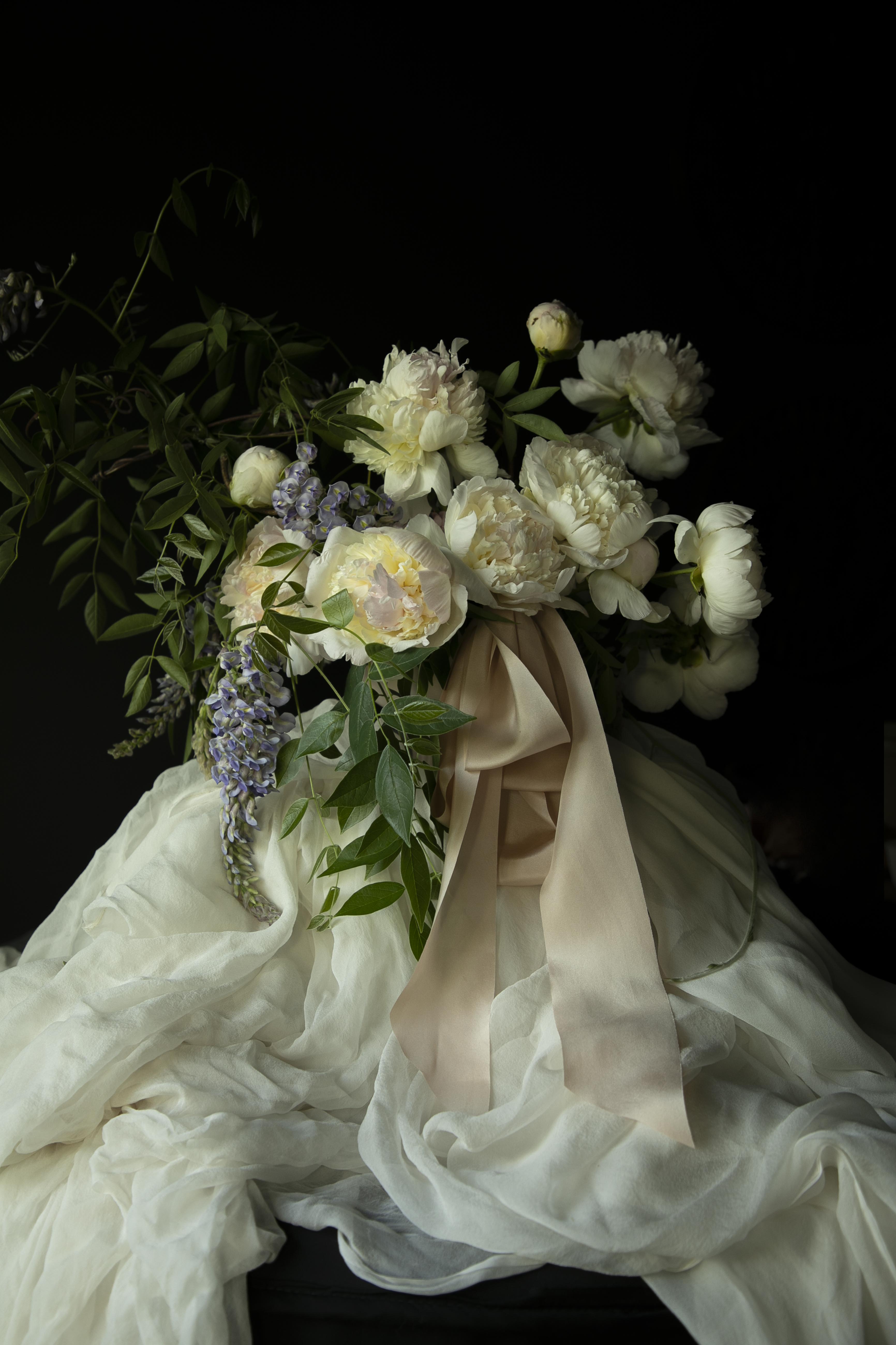 Floral arrangement with white flowers and green leaves draped over a white fabric against a dark background. highlighting the best wedding silk ribbon in blush color.