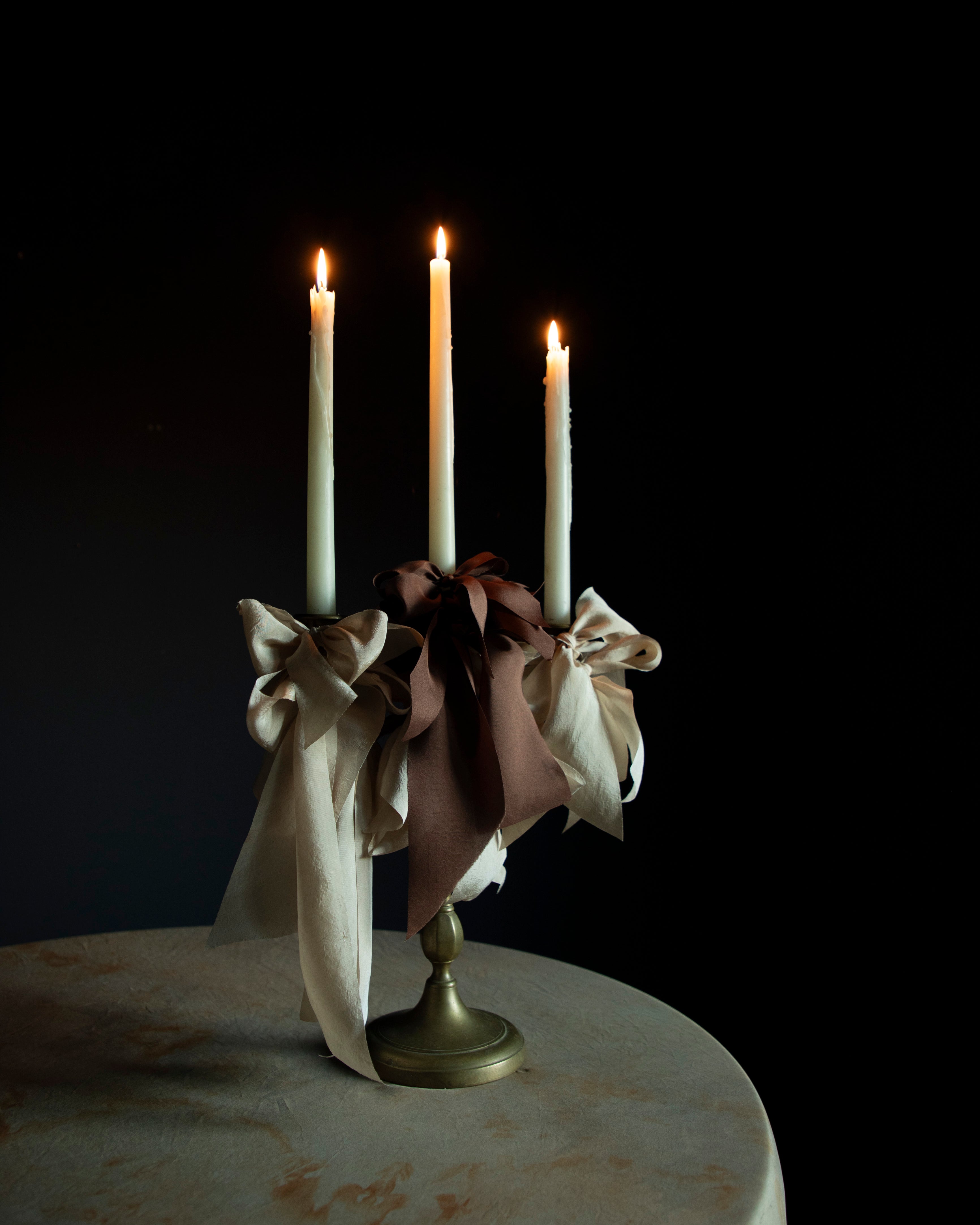 A mocha silk ribbon tied in a bow around a candlestick holder with lit candles, set against a dark background.