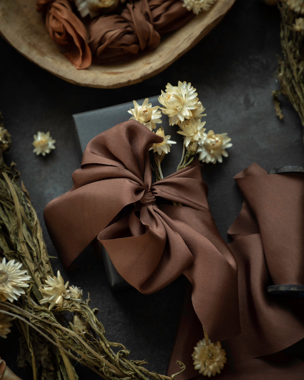 A styled flat-lay of a brown silk ribbon tied in a large bow around a dark box, surrounded by dried flowers and neutral-toned fabric pieces on a dark textured surface.