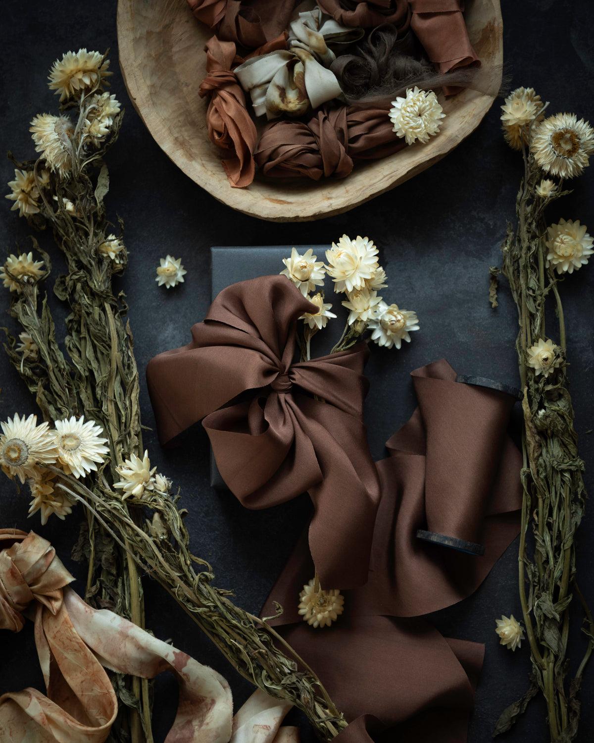 A styled flat-lay of brown silk ribbon or fabric in multiple tied bows, arranged with dried white flowers on a dark textured surface. A wooden bowl at the top contains additional folded brown fabric, and more ribbon and floral props extend across the frame.