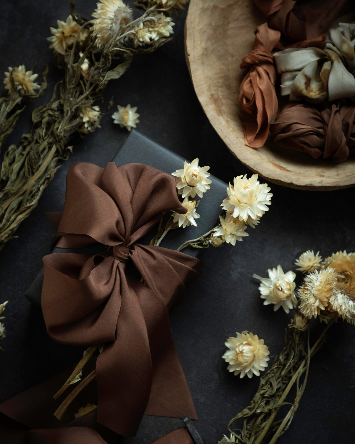 A styled flat-lay featuring a large, glossy dark brown satin ribbon bow on a dark surface, surrounded by dried cream flowers and a wooden bowl filled with more brown and beige ribbons.