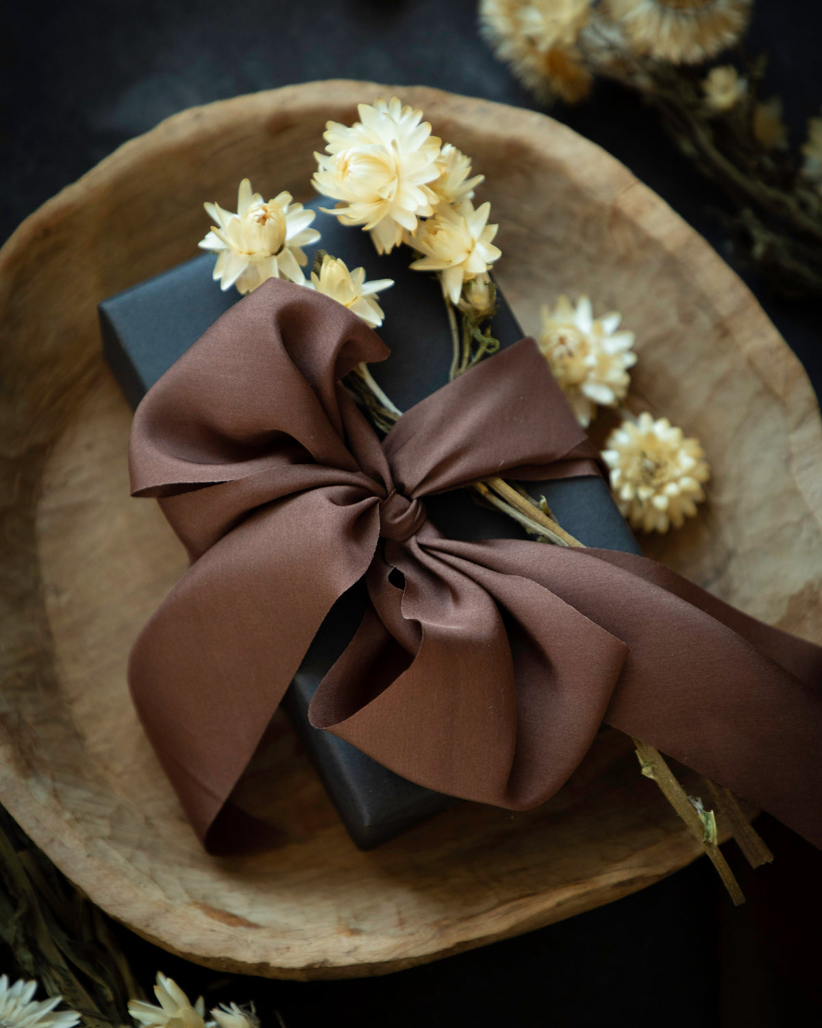 A dark gray gift box wrapped in a large brown satin bow and decorated with dried cream flowers, presented on a rustic wooden plate against a dark background.