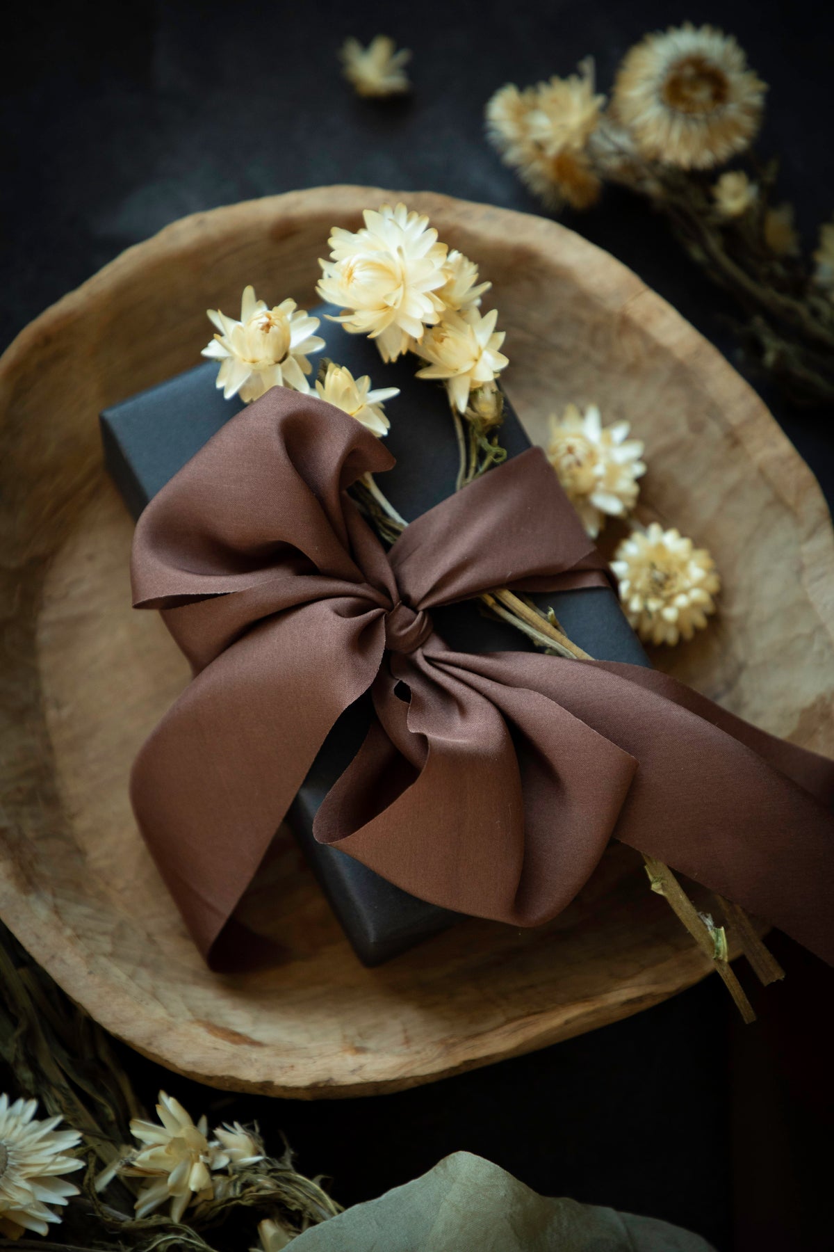 A dark gray gift box wrapped in a large brown satin bow and decorated with dried cream flowers, presented on a rustic wooden plate against a dark background.