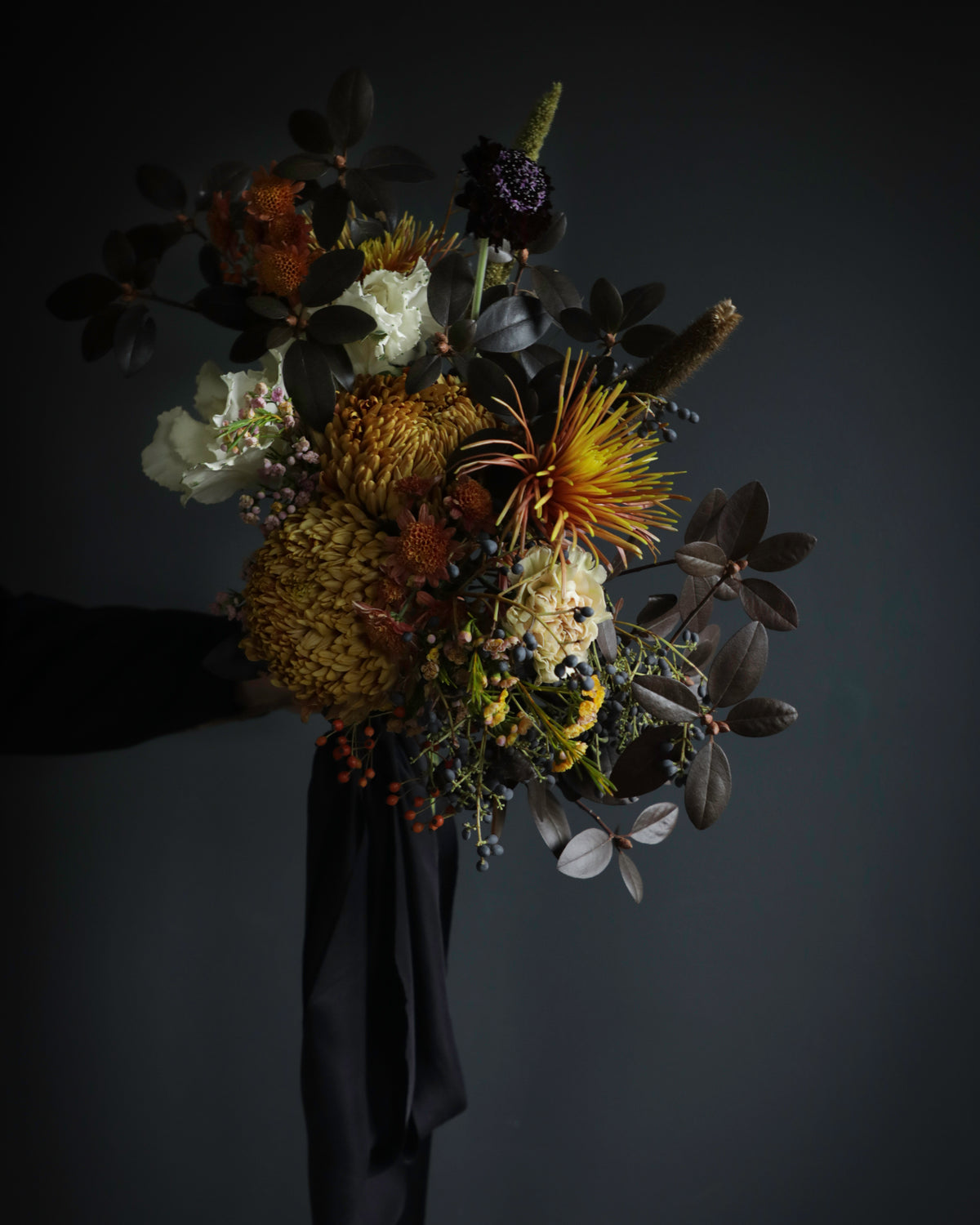A rustic flower bouquet featuring a mix of orange, yellow, white, and dark brown botanicals with a long black ribbon, photographed against a dark gray background.